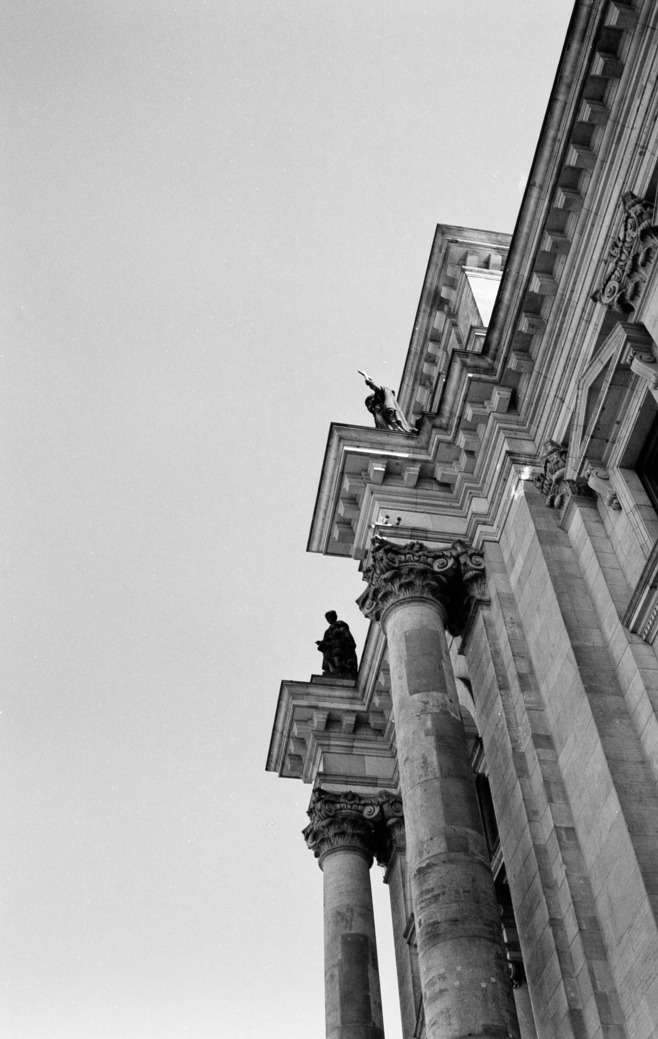 Black and white photo of a historic building's ornate exterior, including tall columns and detailed sculptures on top, viewed from a low angle against the sky.