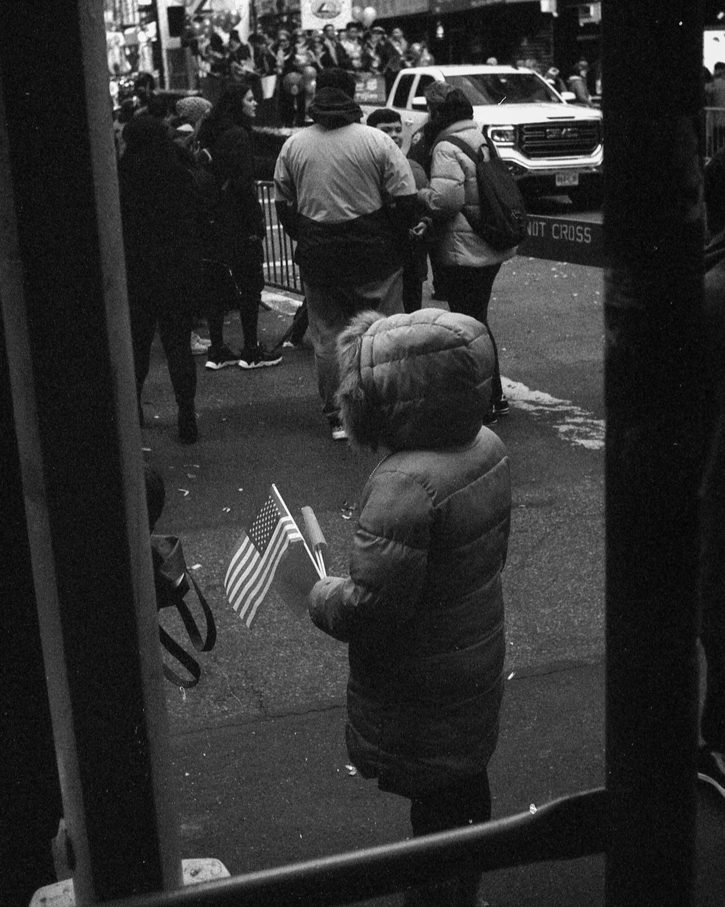 A group of people, including children and adults, are gathered outside an urban store or shopping area, with some individuals wearing winter jackets. There are American flags on a chair, and a vehicle is visible in the background. The scene appears t
