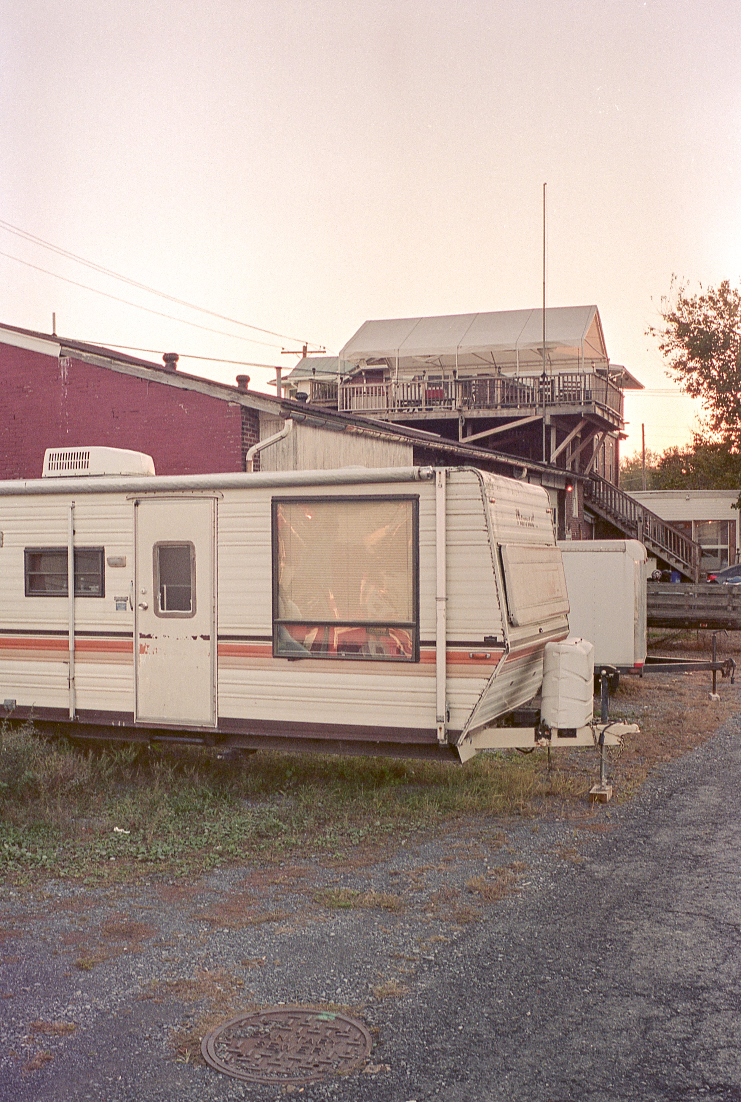 A vintage white travel trailer with an orange stripe parked on a gravel lot, with a red brick building and wooden deck in the background.