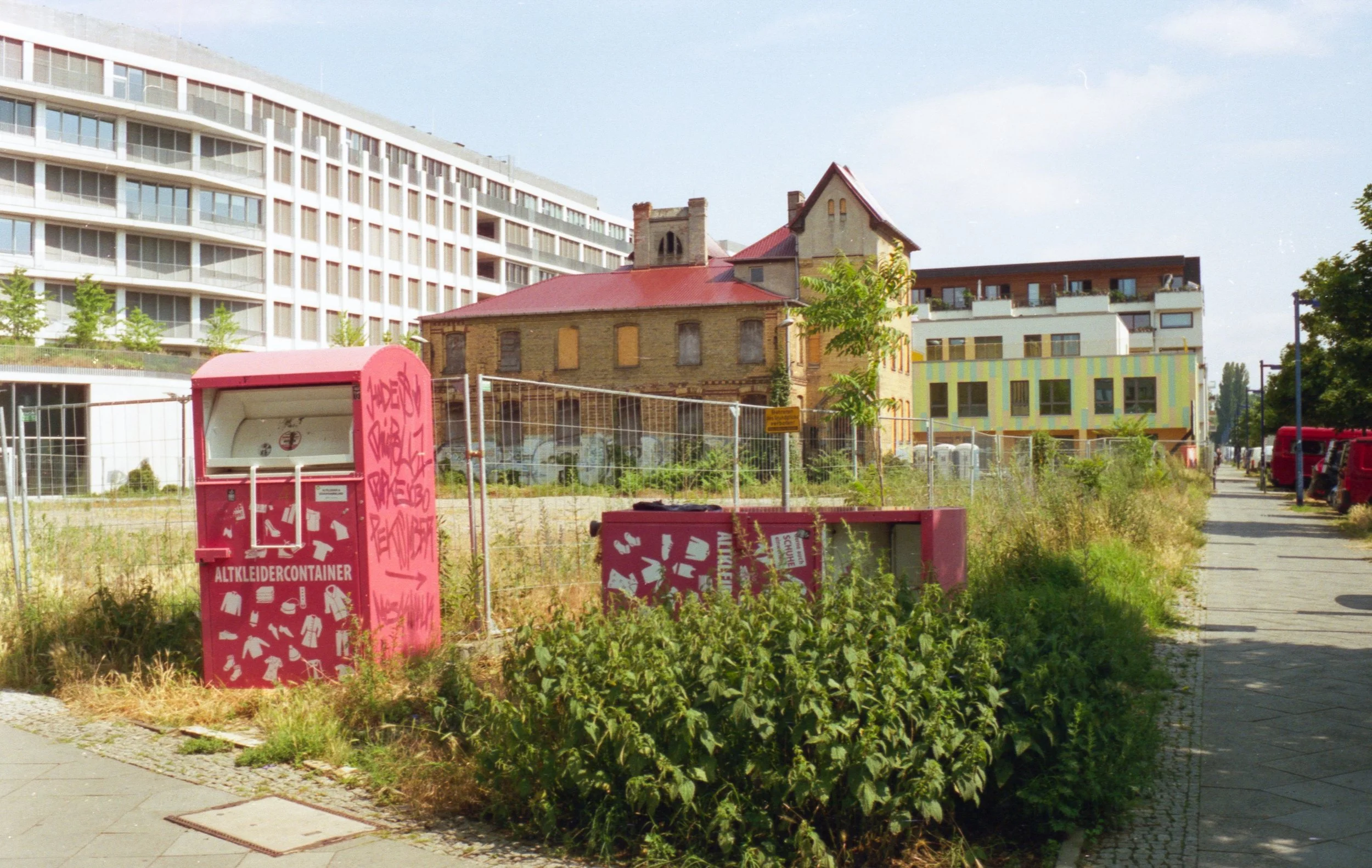 A construction site with overgrown plants and graffiti-covered pink recycling bins in the foreground, an old abandoned brick building with boarded-up windows in the middle ground, and modern white residential buildings in the background.