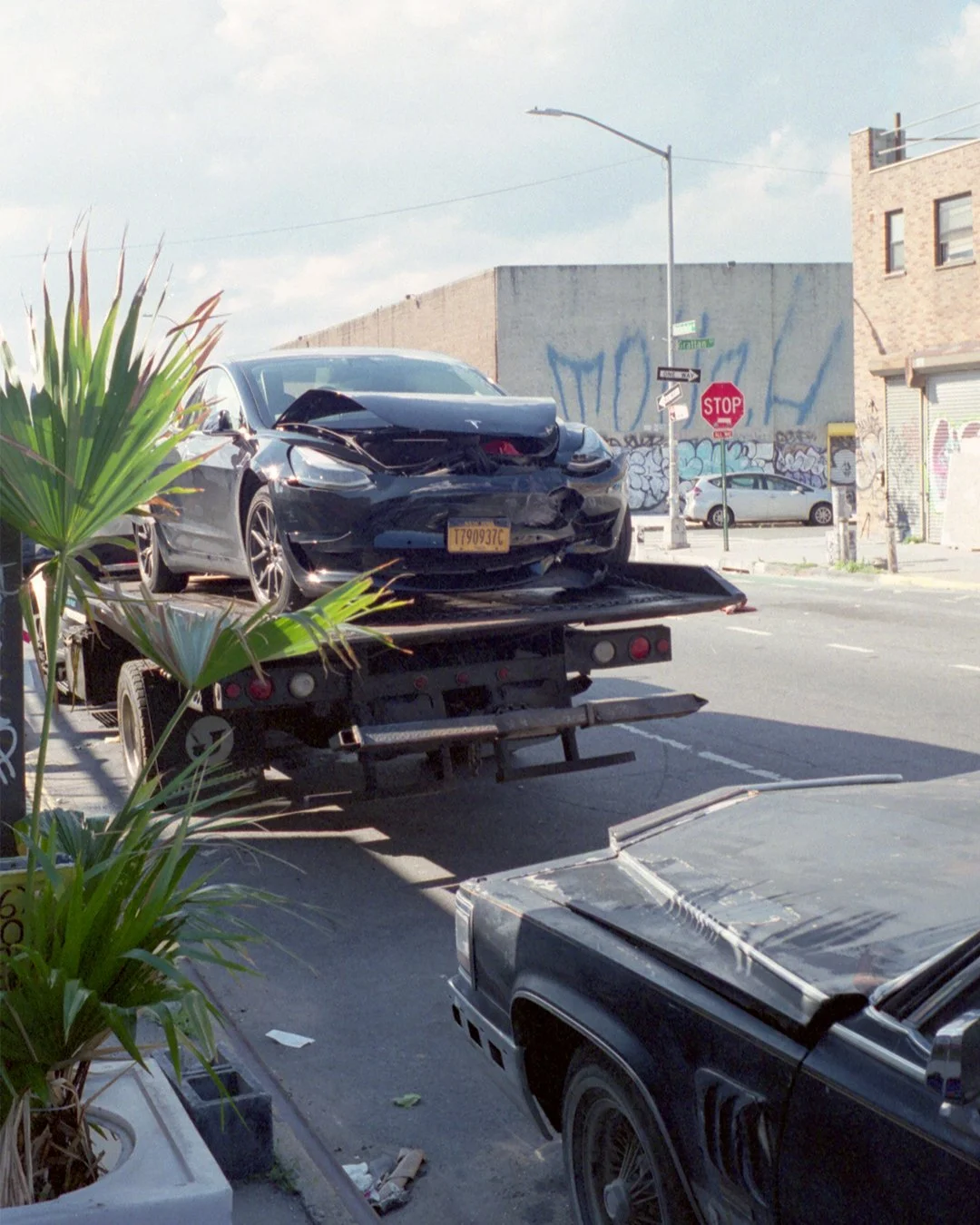 A damaged black Tesla car on a flatbed tow truck at an urban street corner with graffiti on walls and a stop sign.