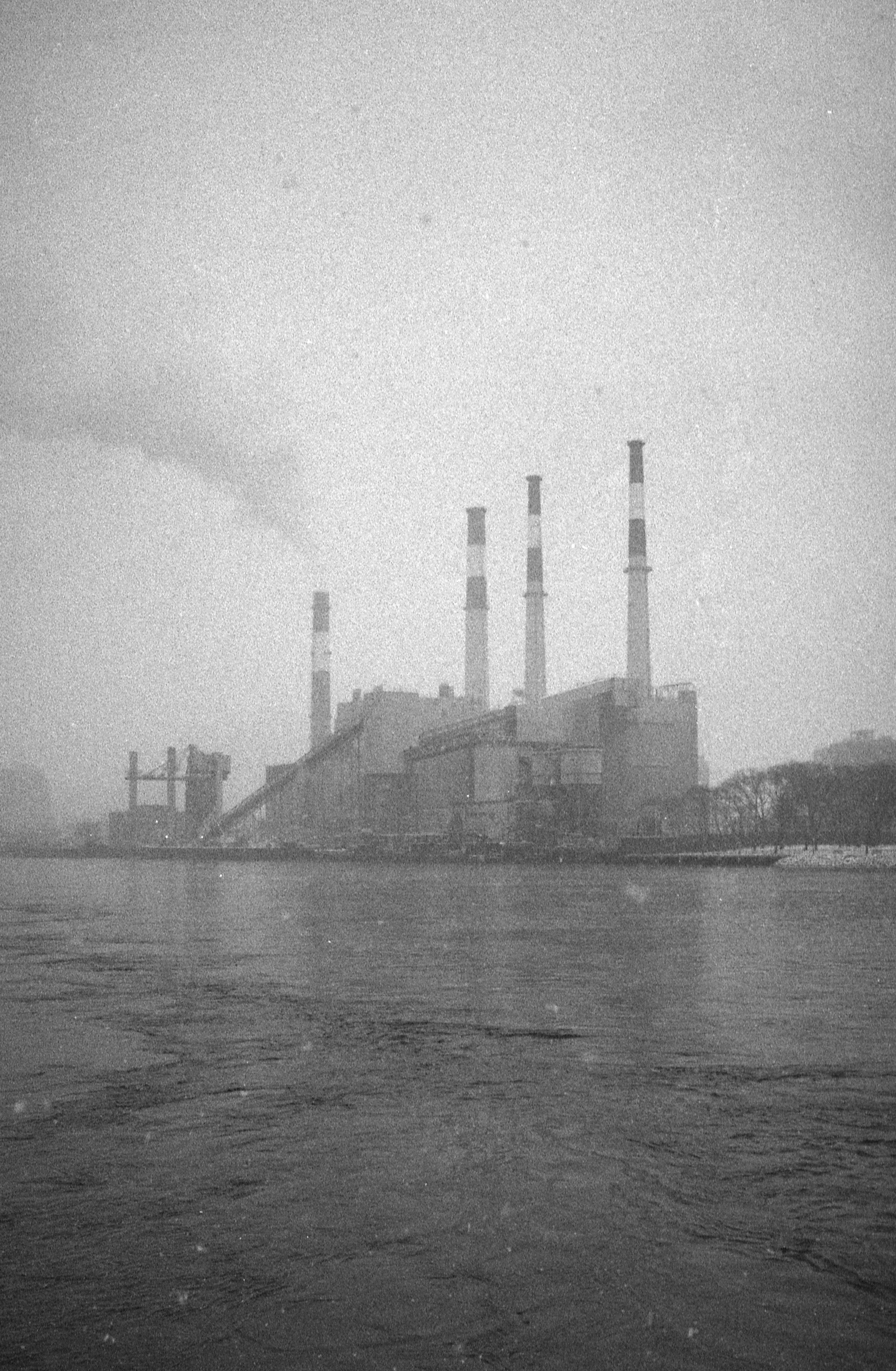 Industrial factory with tall smokestacks emitting smoke, situated near a body of water, under a cloudy sky.