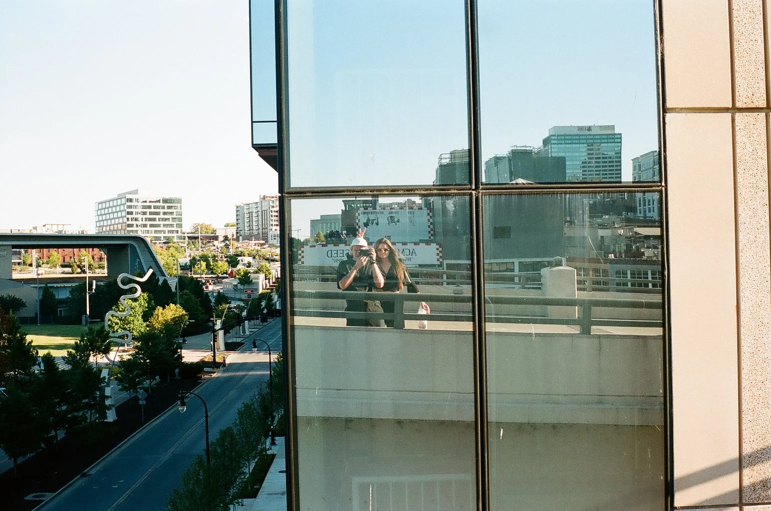 Two women are taking a photo of each other in a reflection on a glass window of a building in an urban area. The cityscape includes various modern buildings, trees, and a street below.