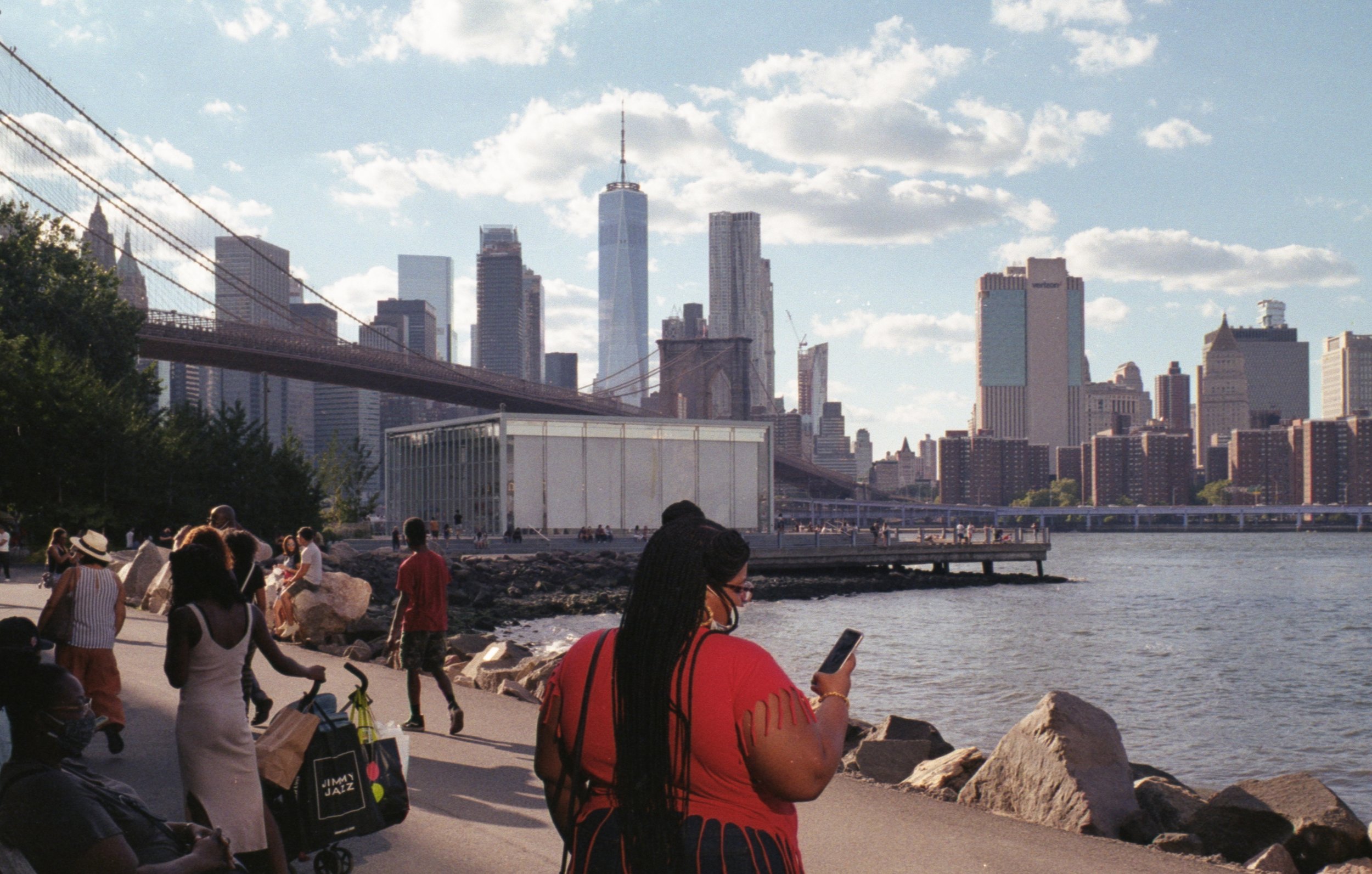 People sitting and walking along the waterfront in New York City with the Brooklyn Bridge and Manhattan skyline in the background, including One World Trade Center and various skyscrapers, under a partly cloudy sky.