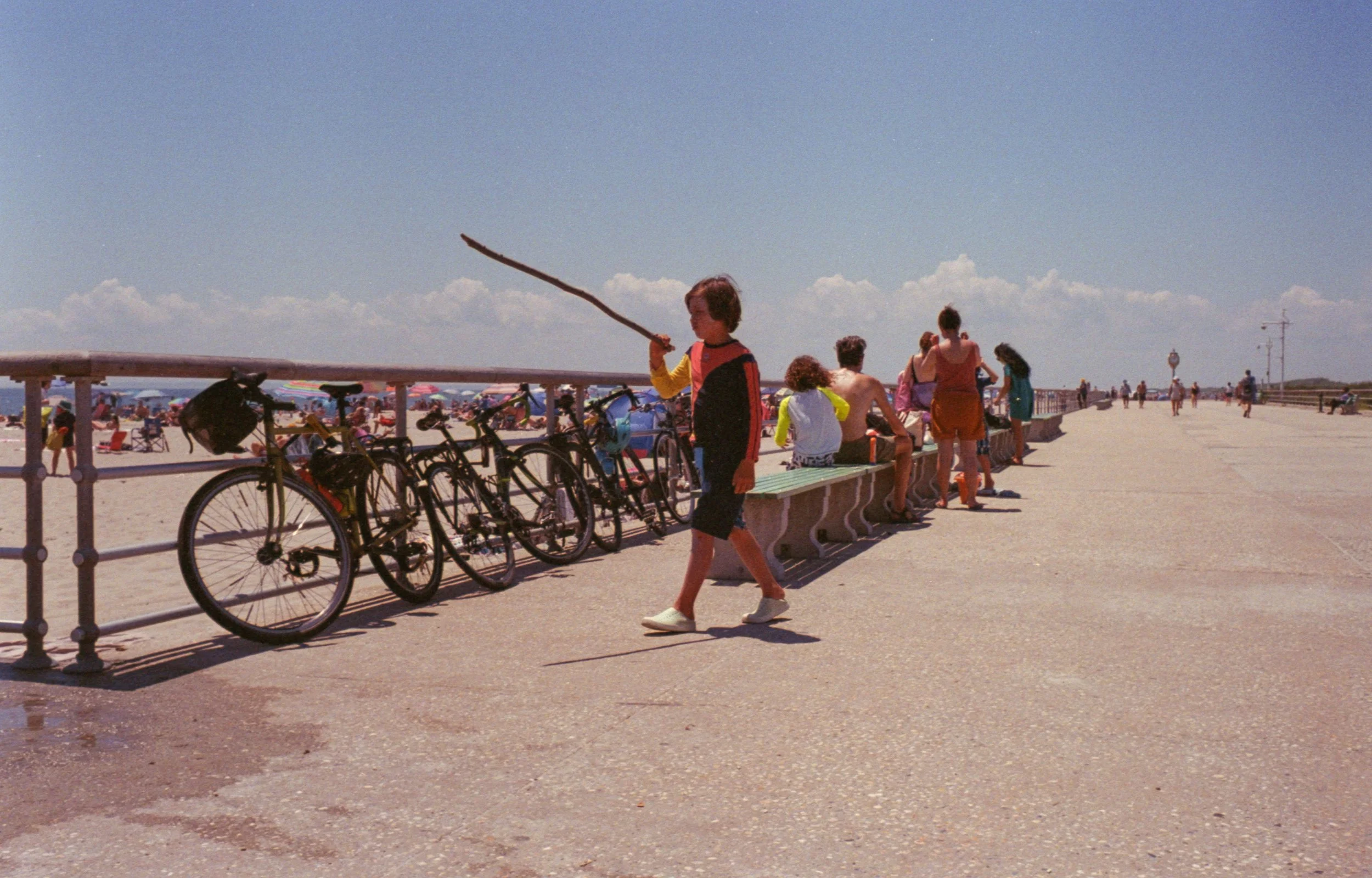 People at the beach promenade with bicycles, umbrellas, and seating area, under a clear blue sky.
