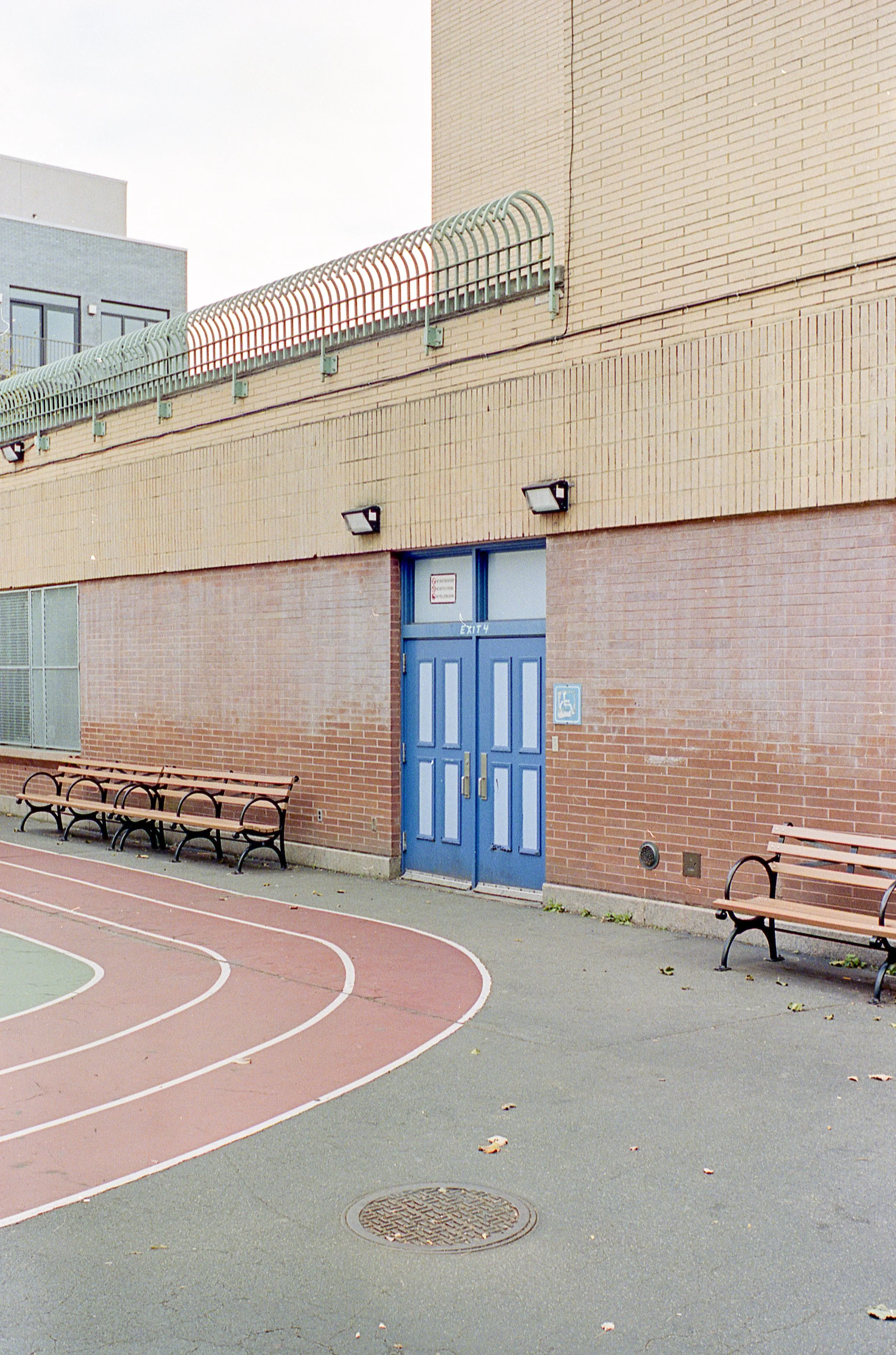 Empty outdoor running track with benches, a brick building with blue double doors, and a high wall with a balcony on top.