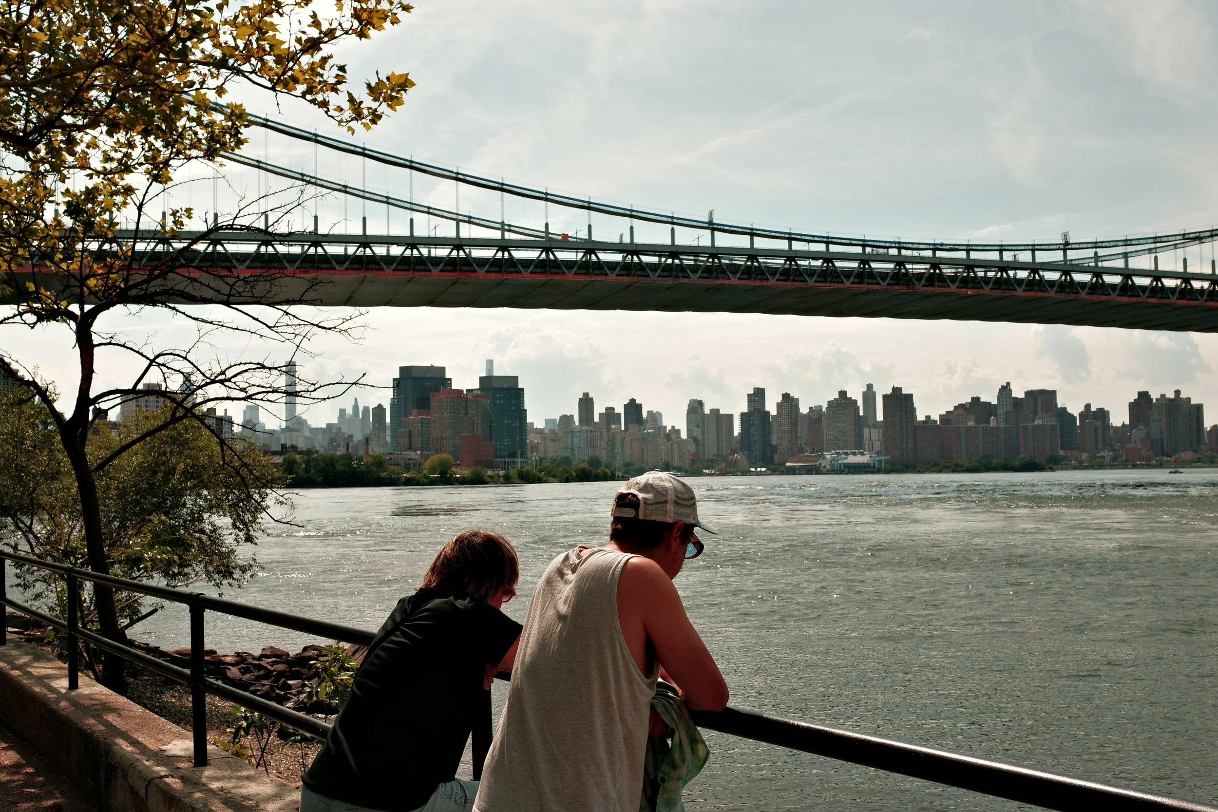 People sitting by a river with a city skyline and a bridge in the background.