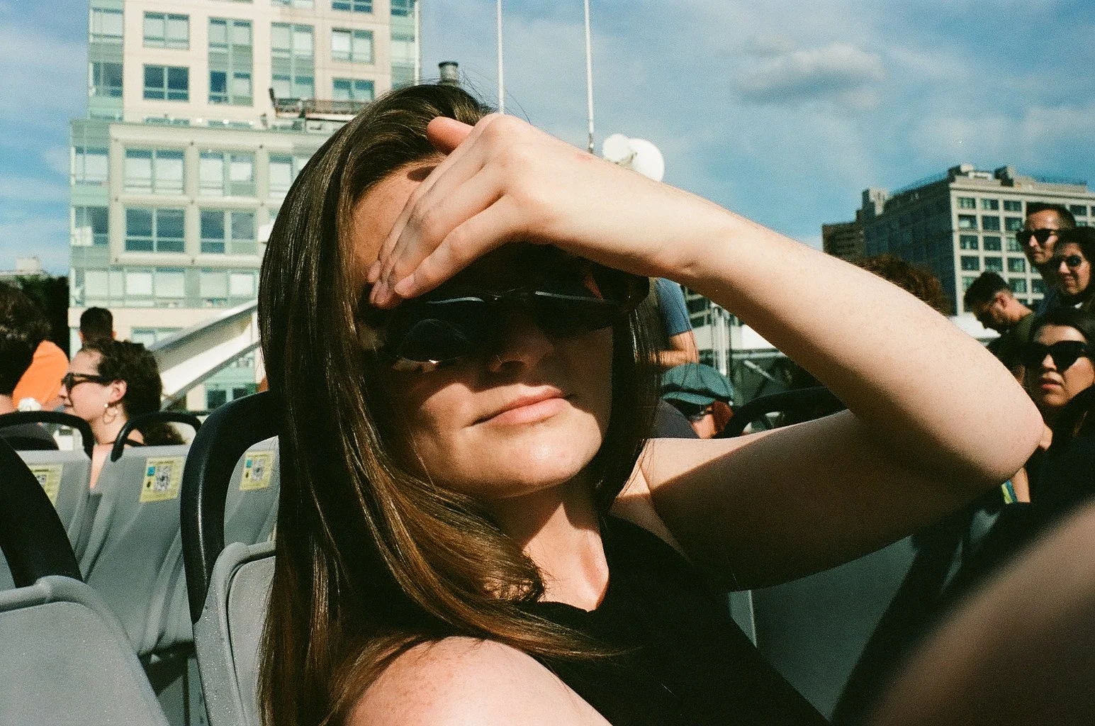 A woman with dark hair wearing sunglasses is sitting outdoors in a bus or tour vehicle, shielding her eyes from the sun with her hand. In the background, there are other people, some wearing sunglasses, and city buildings under a partly cloudy sky.