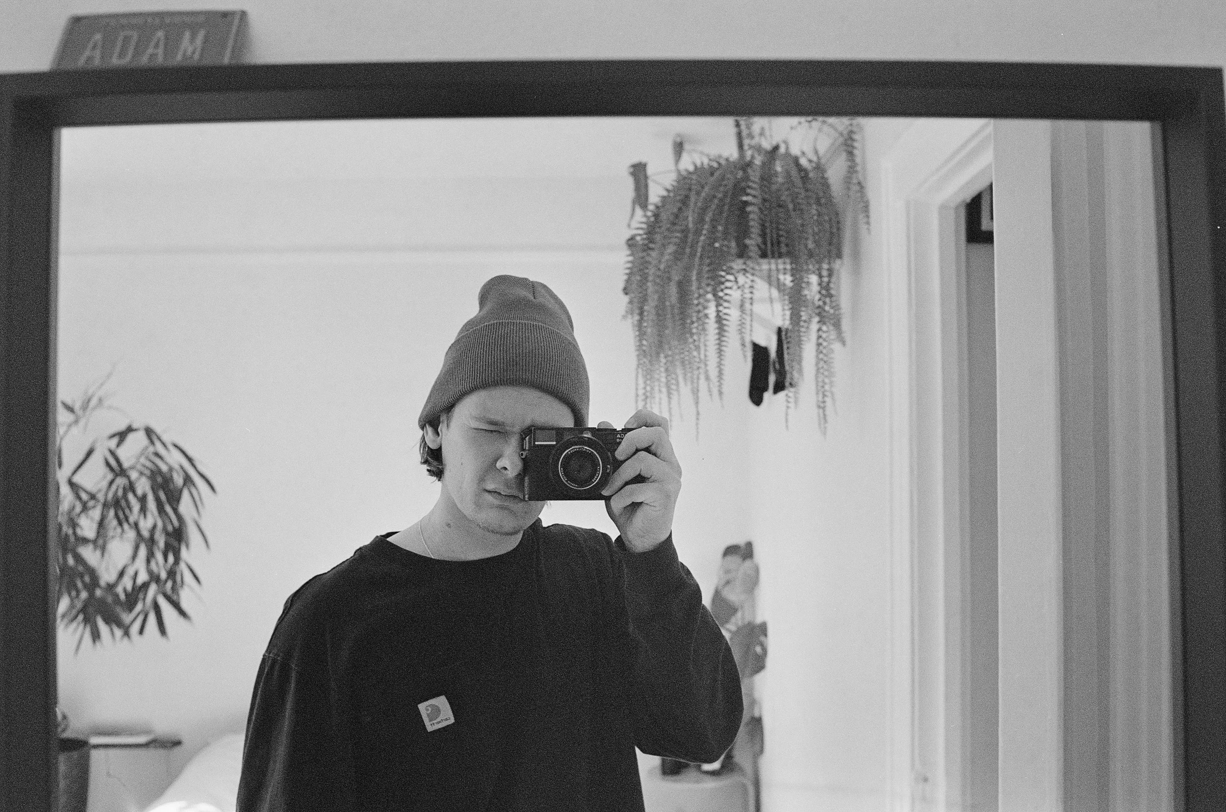 A person wearing a beanie taking a selfie in a mirror, with a plant and clothes hanging in the background.