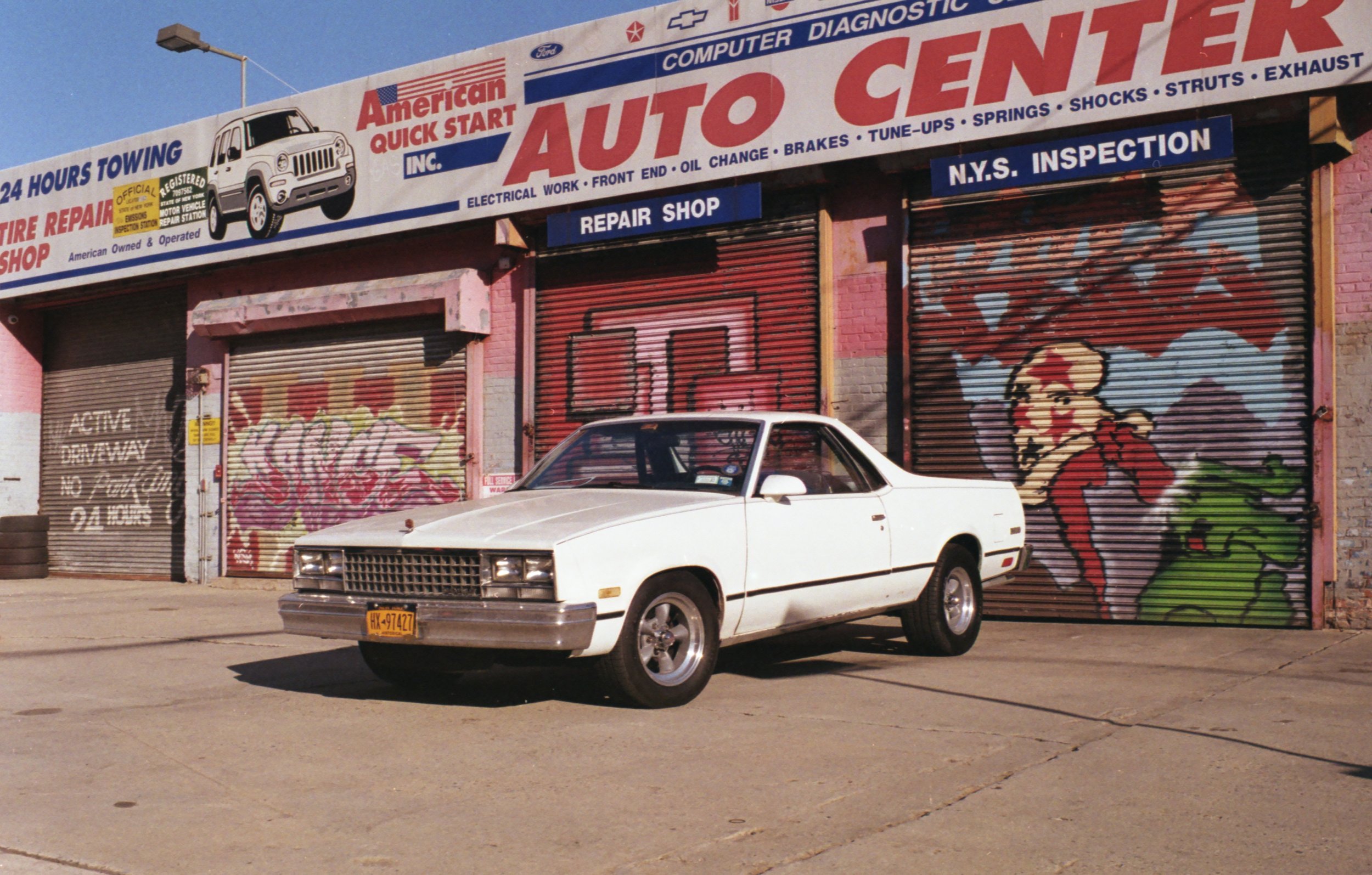 A white vintage car parked in front of a closed roller shutter with graffiti on it, in front of a colorful storefront with signage for an auto repair shop.