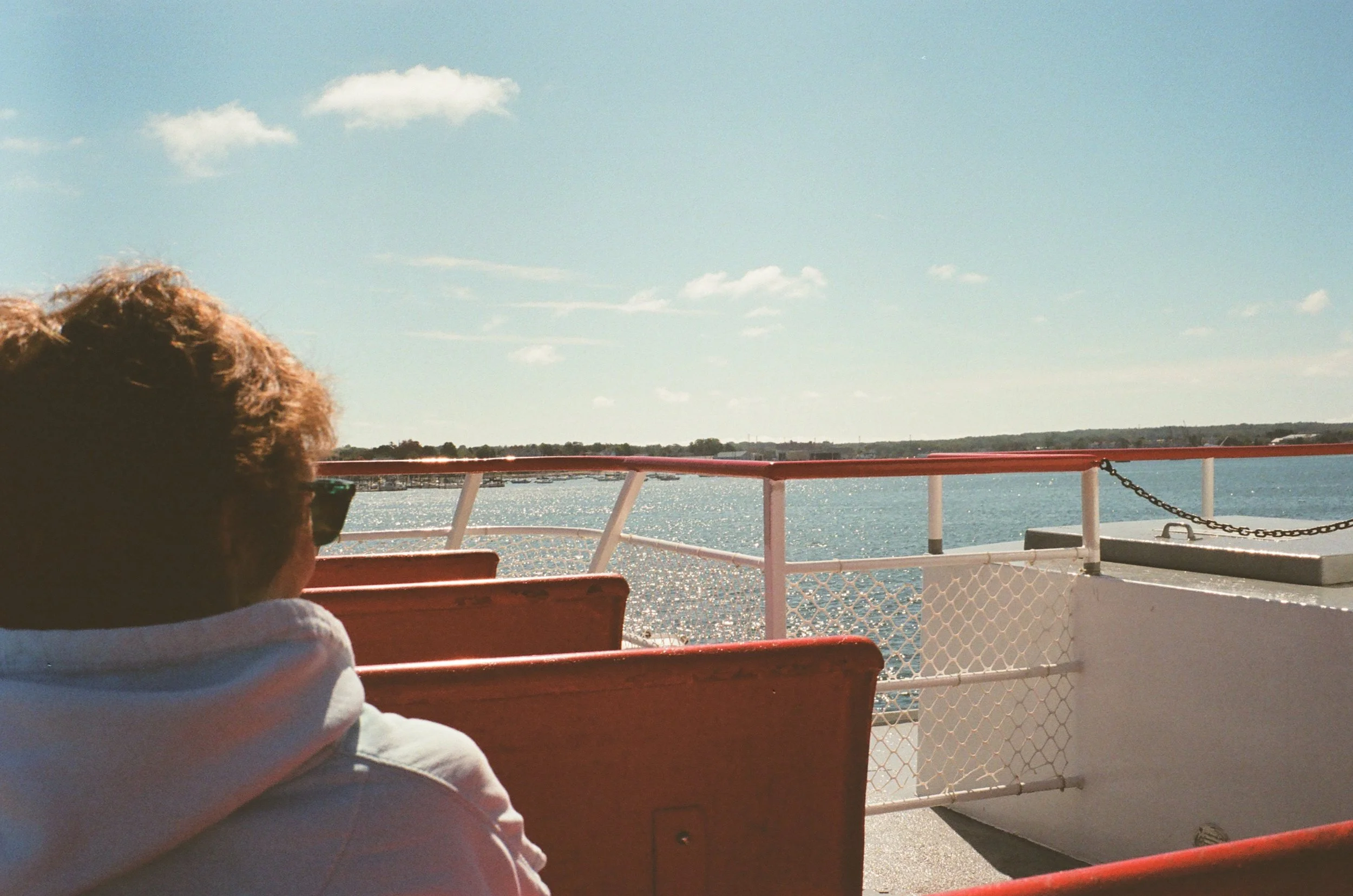 A person with curly hair and sunglasses sitting on a boat, with water and partly cloudy sky in the background.