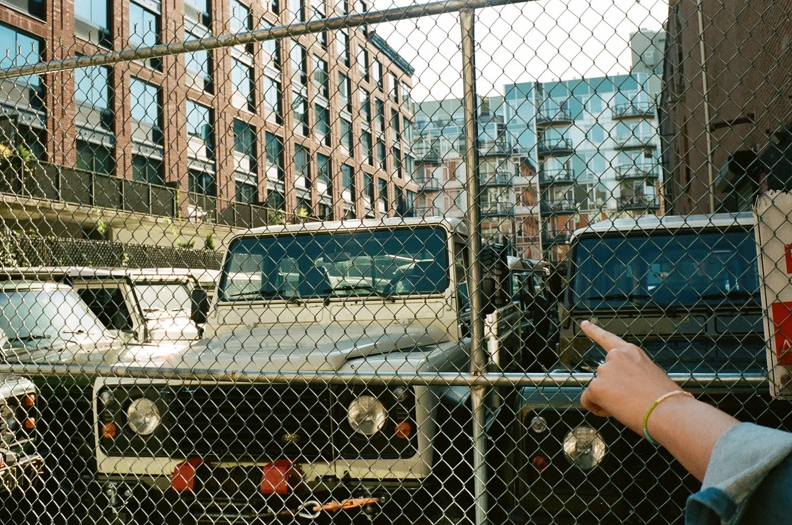 A person's hand pointing at vintage trucks parked behind a chain-link fence, with colorful apartment buildings in the background.