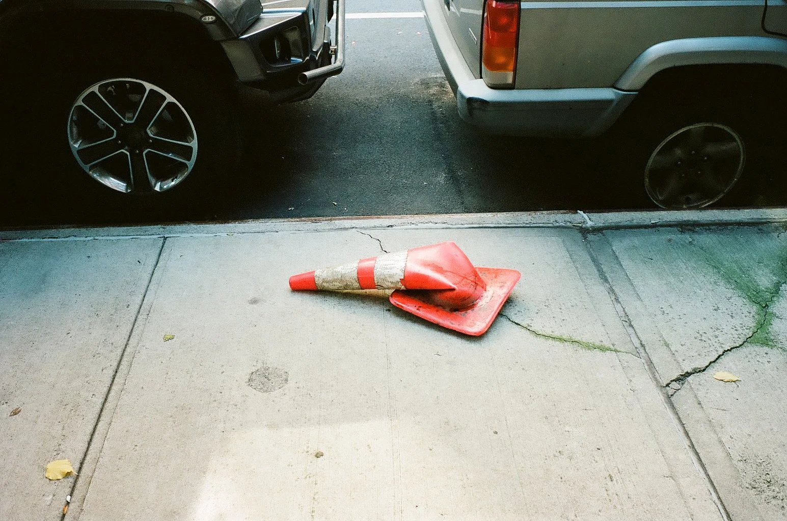 A fallen orange and white traffic cone on a concrete sidewalk outside, with cracked pavement and scattered leaves.