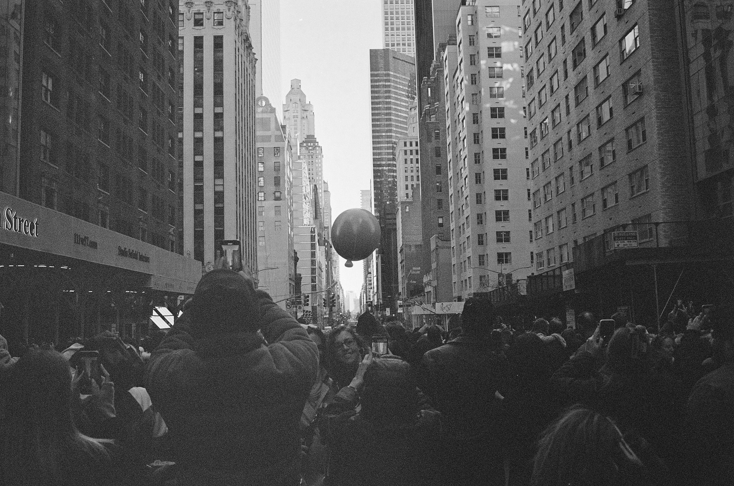 Crowded city street filled with pedestrians, tall buildings lining both sides, and a hot air balloon floating above.