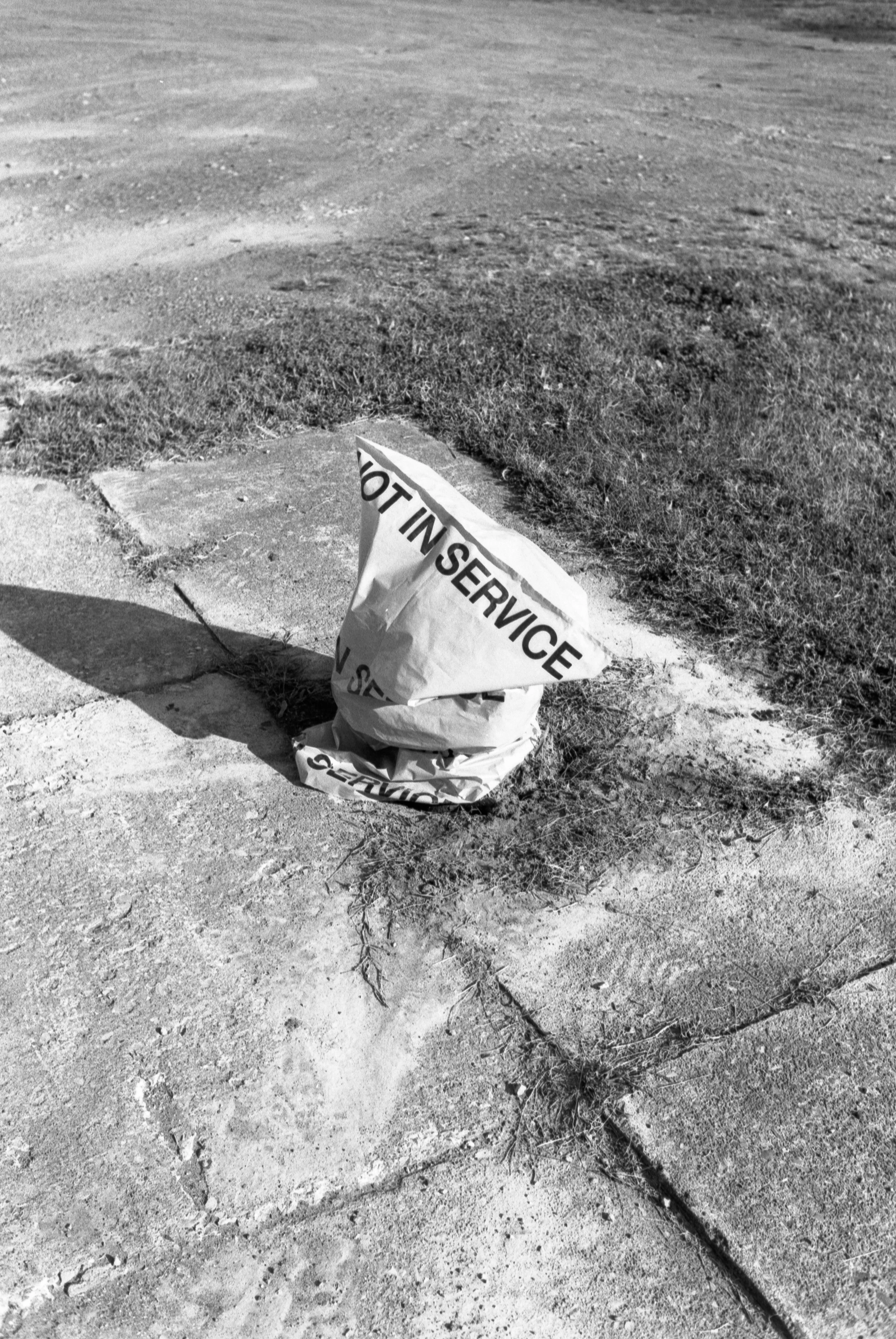 A crumpled paper bag with the words 'DO NOT IN SERVICE' written on it, placed on a sidewalk curb next to a patch of grass and dirt.