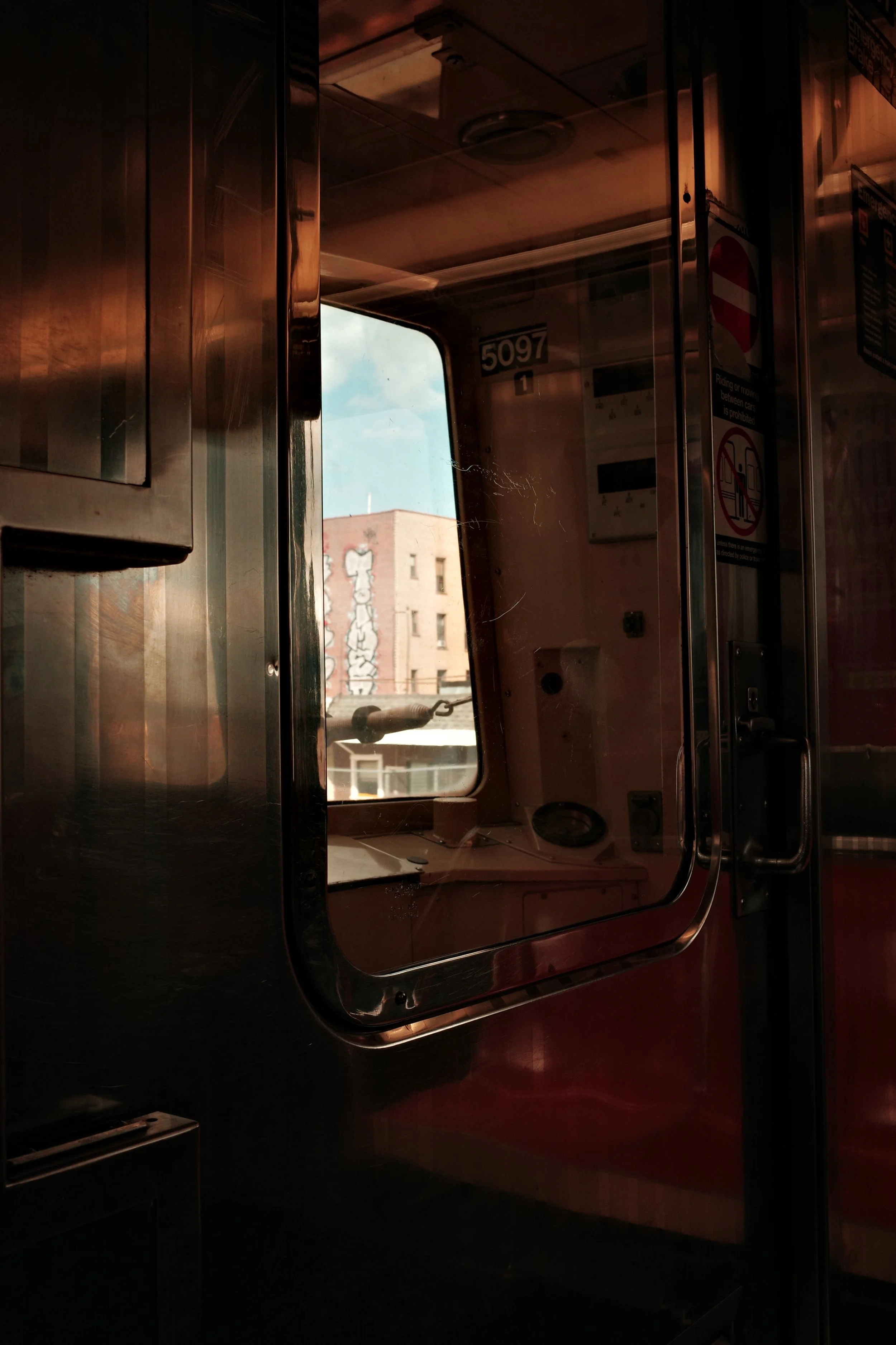 Inside a train car with reflective metal surfaces and a window showing buildings and a blue sky outside.