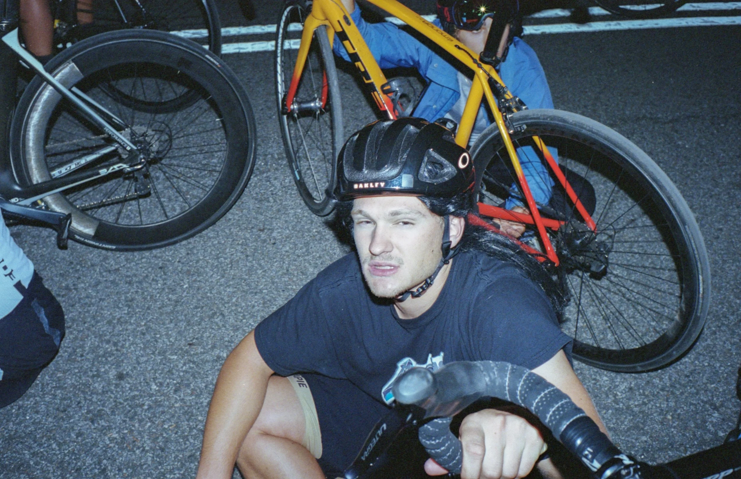 A man in a black cycling shirt and helmet lies on the asphalt ground, holding a black mountain bike. He is being attended to by another cyclist, partially visible on the left, and surrounded by cycling gear and equipment, suggesting an outdoor cyclin