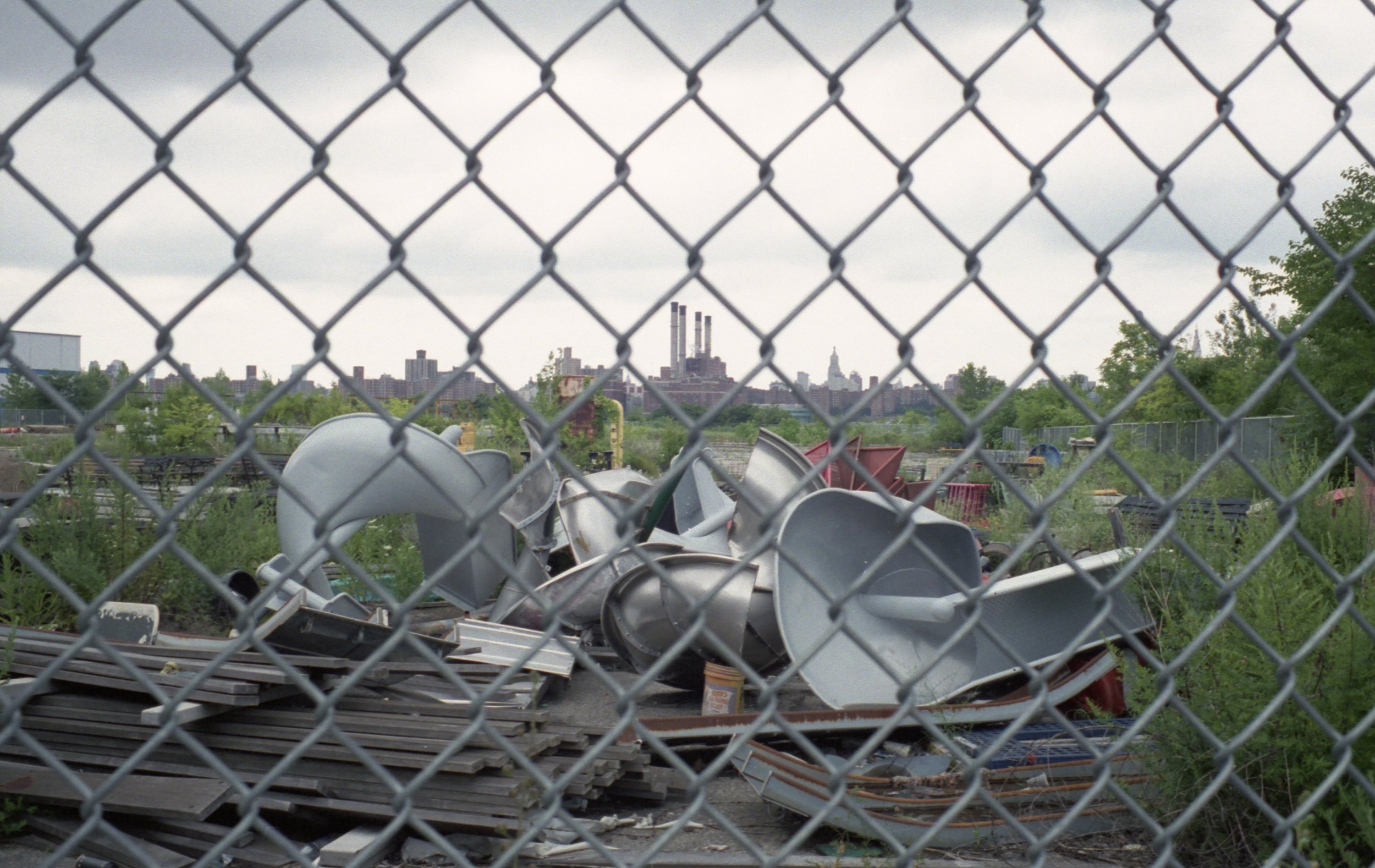 A view of an industrial area with a chain-link fence in the foreground, cluttered with scrap metal and construction debris. In the background, there are city buildings and a cloudy sky.