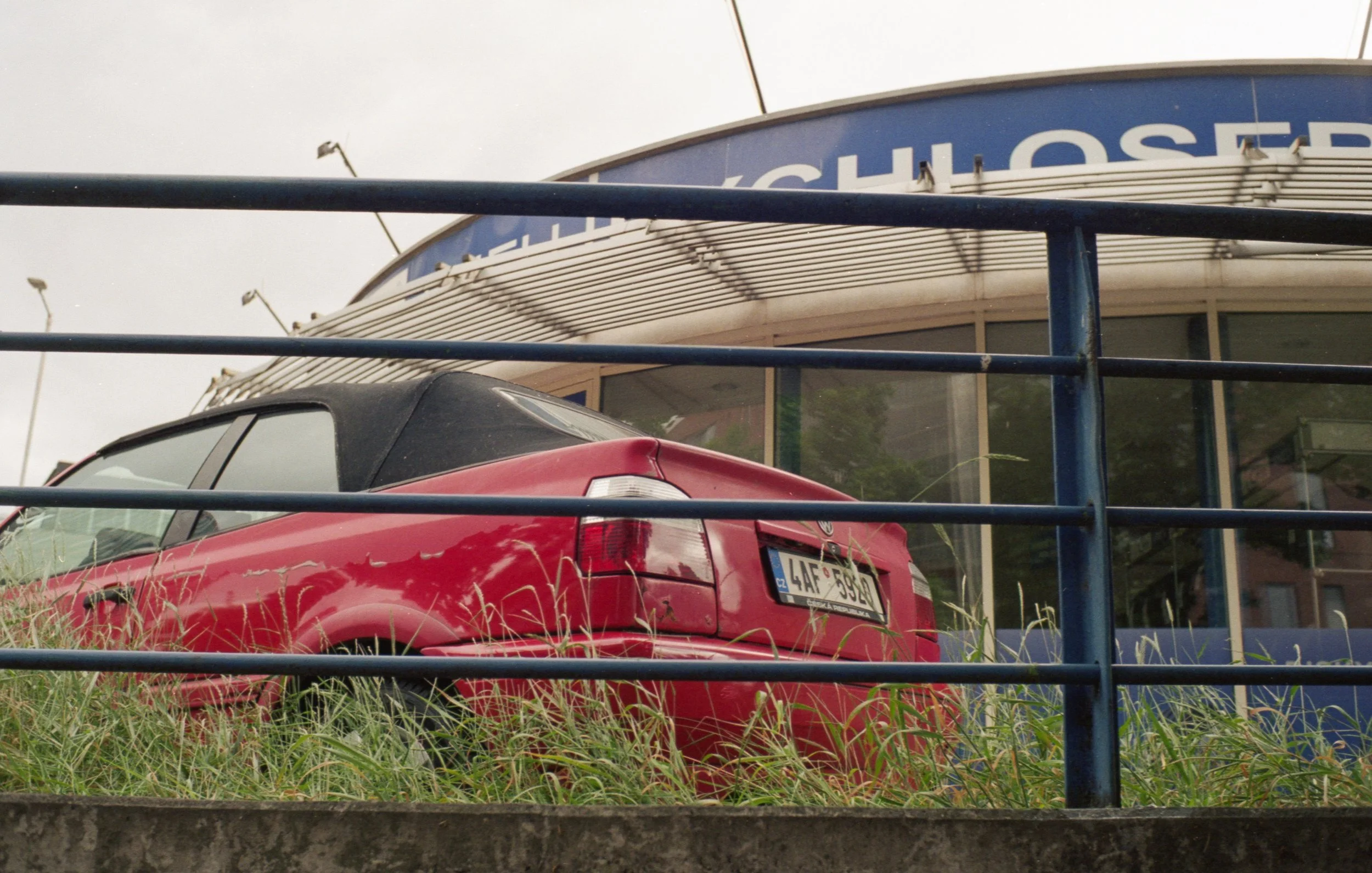 Red car parked behind a blue metal fence in front of a building with a sign reading 'CHLOR...'