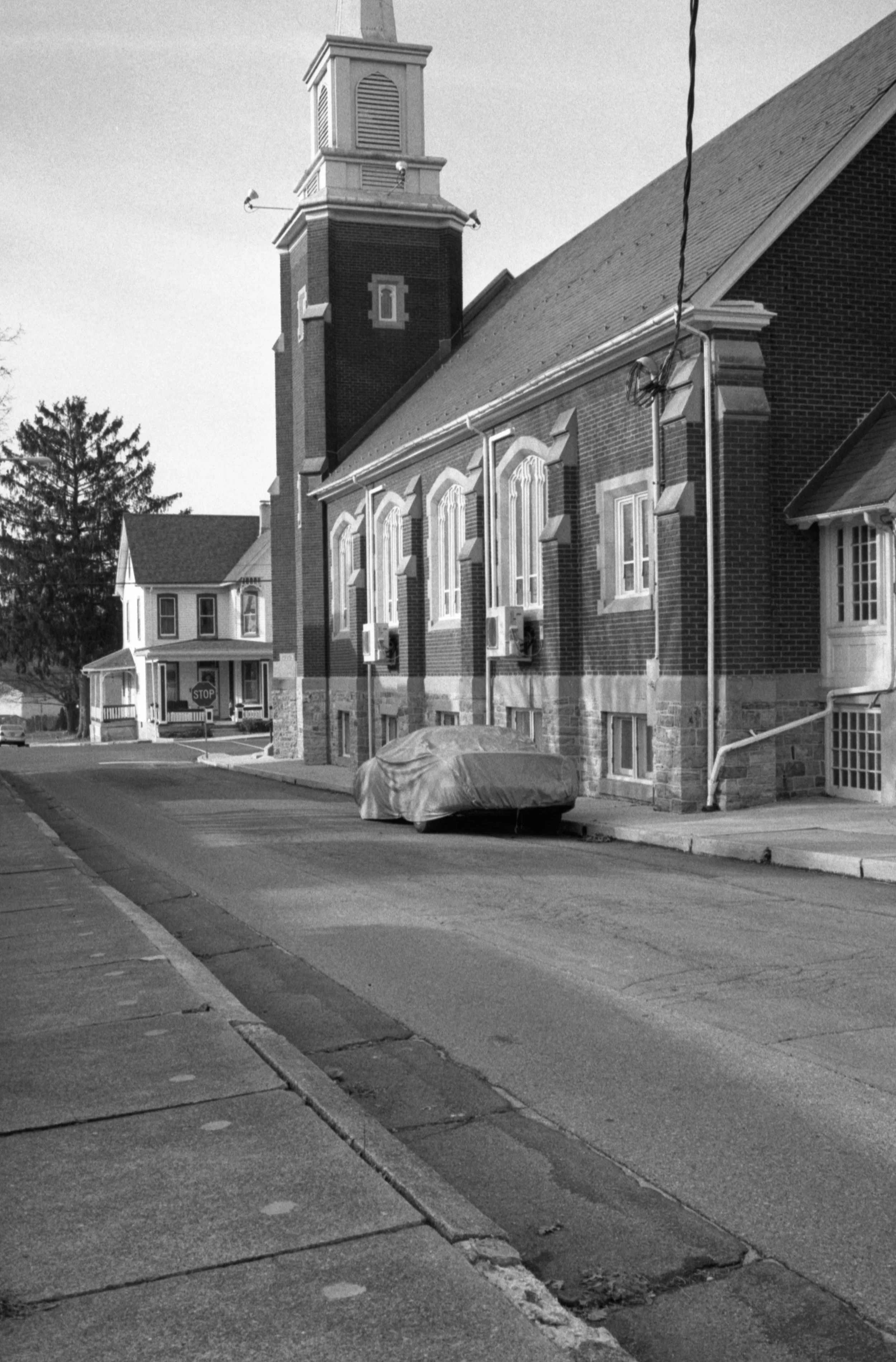 A black and white image of a street view showing a brick church with a tall steeple, multiple windows, and air conditioning units on the building's exterior. A covered car is parked on the sidewalk, and there is a stop sign at the corner. In the back