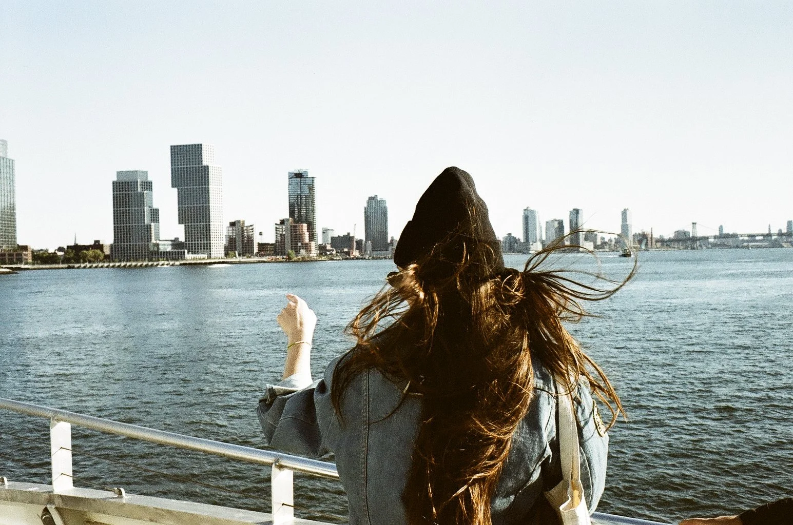 A woman with long hair, wearing a hoodie and denim jacket, standing on a boat or dock near water, facing away from the camera, with a city skyline in the background.