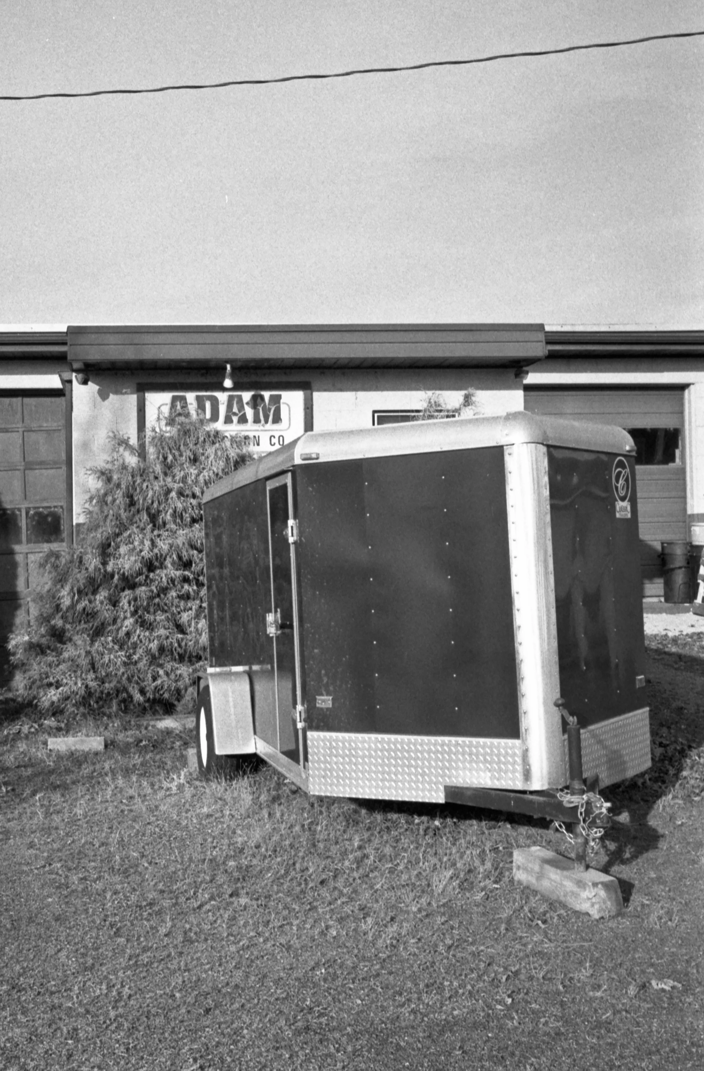 Black and white photo of a trailer parked on grass in front of a building with a sign that reads 'ADAM'. The trailer is attached to a hitch, and there is a wooded shrub next to the trailer, with a small brick and block on the ground nearby.