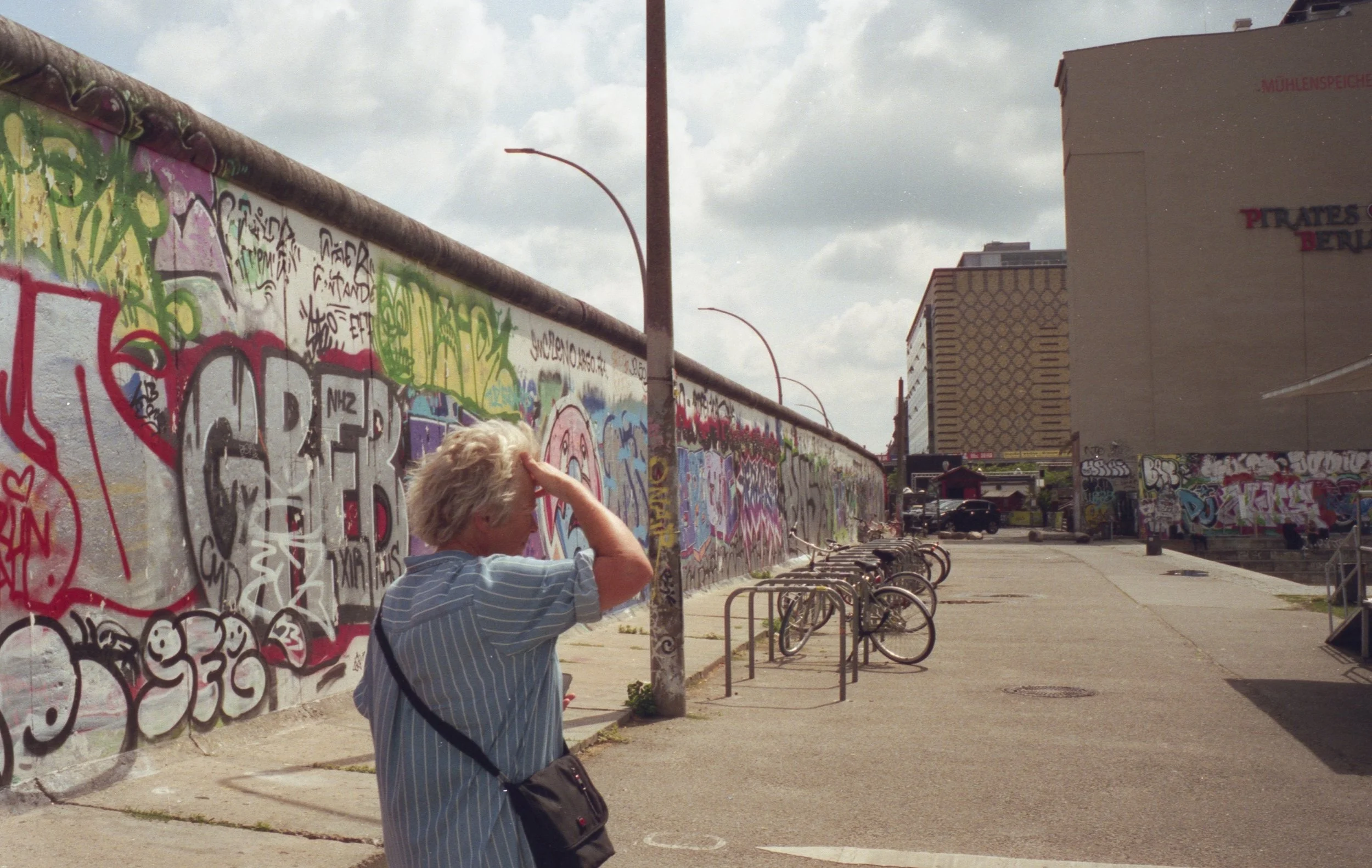 An older woman with white hair wearing a blue striped shirt and a shoulder bag is standing outside near a graffiti-covered wall, touching her head and looking away. There are several bicycles parked along a rack, and a large building in the backgroun