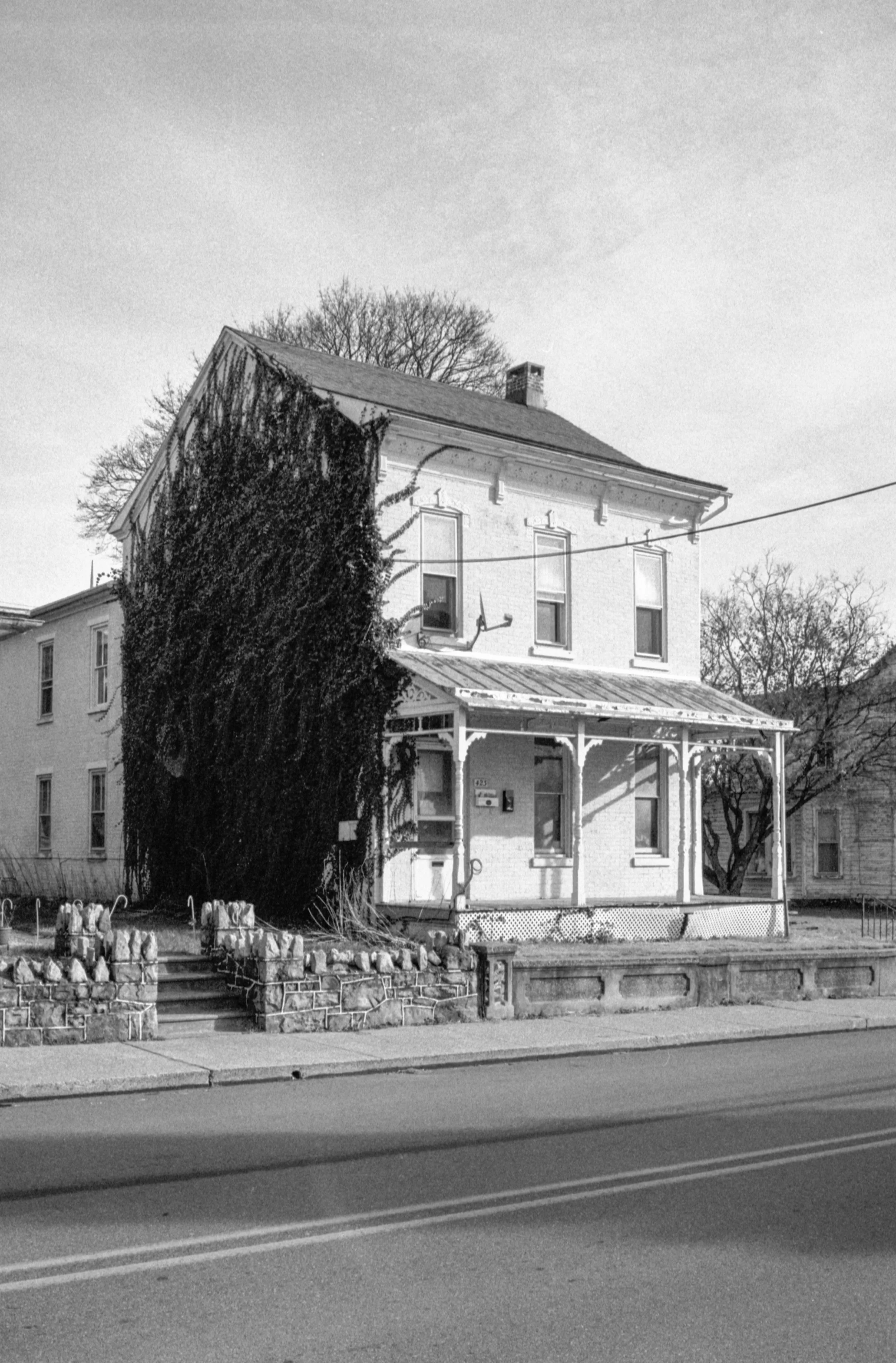Black and white photograph of a two-story house with a front porch and a large ivy-covered tree on the left side. The house has three tall windows on the second floor and a chimney on the roof. There is a stone fence with steps in front of the house 