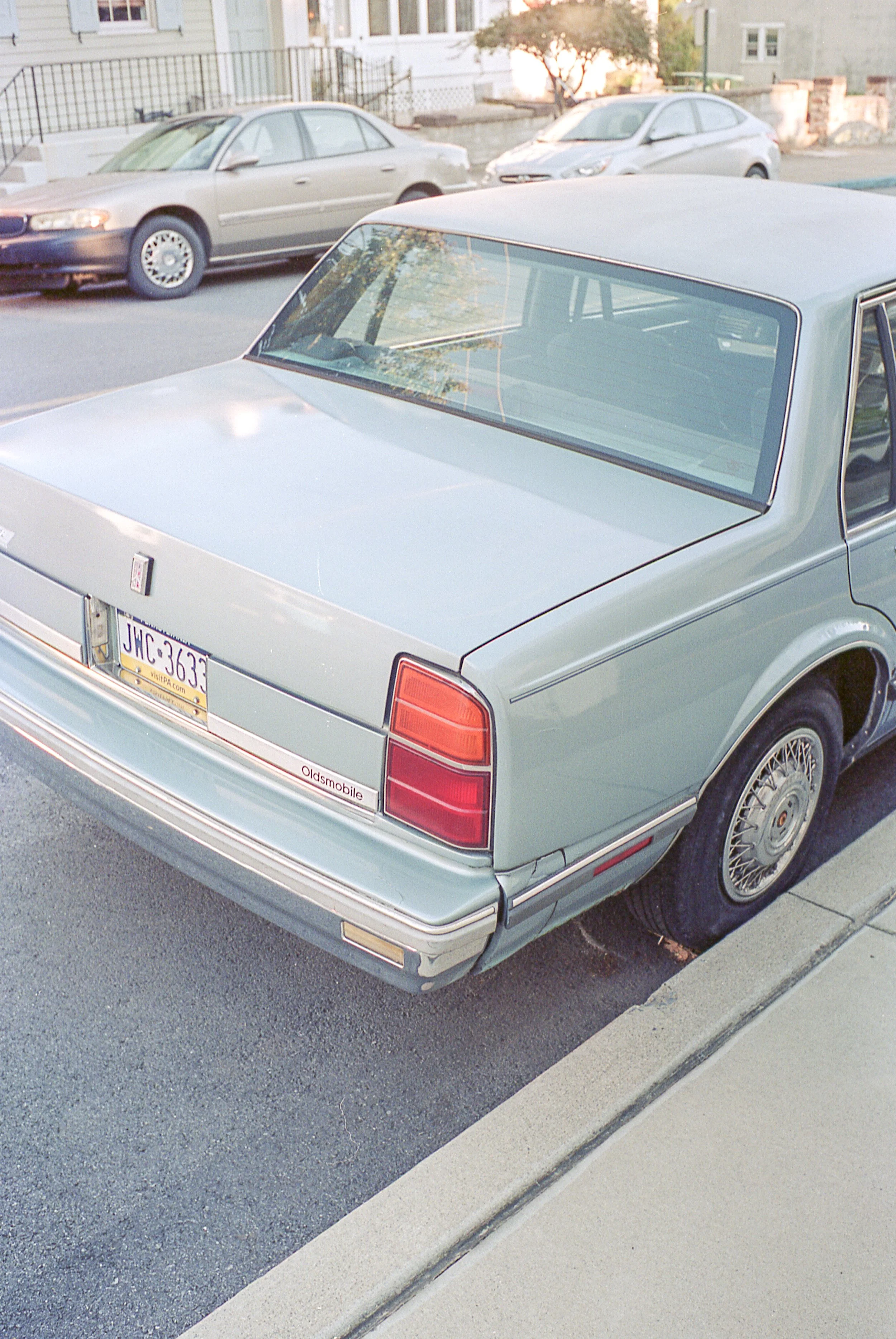 Rear view of a vintage silver Oldsmobile car parked on the street in front of a sidewalk, with other cars and residential buildings in the background.