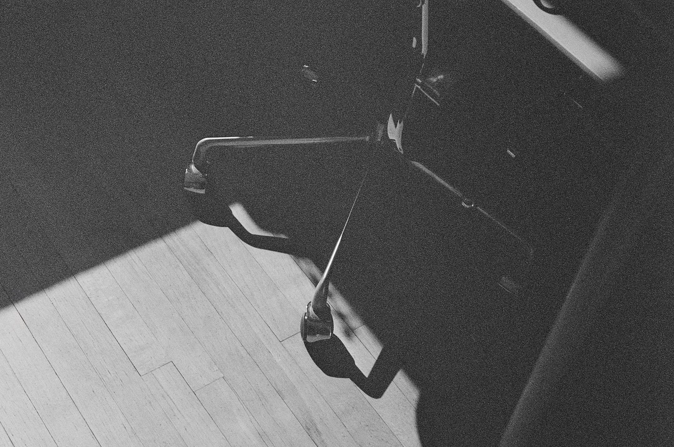 Black and white photo of a closed piano with a stool in front of it, in a room with light-colored wooden floor.