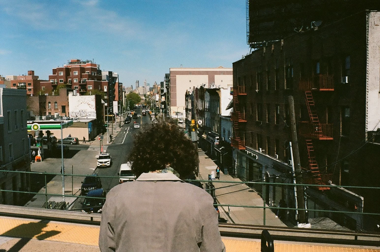 Person with curly hair and gray jacket looking out over urban street from rooftop or balcony, city buildings, parked cars, and clear sky visible.