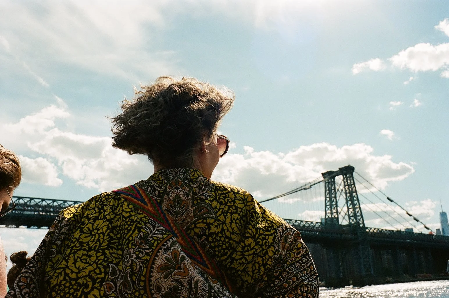 Woman with curly hair and sunglasses wearing a patterned yellow and black shirt, standing near a bridge under partly cloudy sky, with water in the foreground.