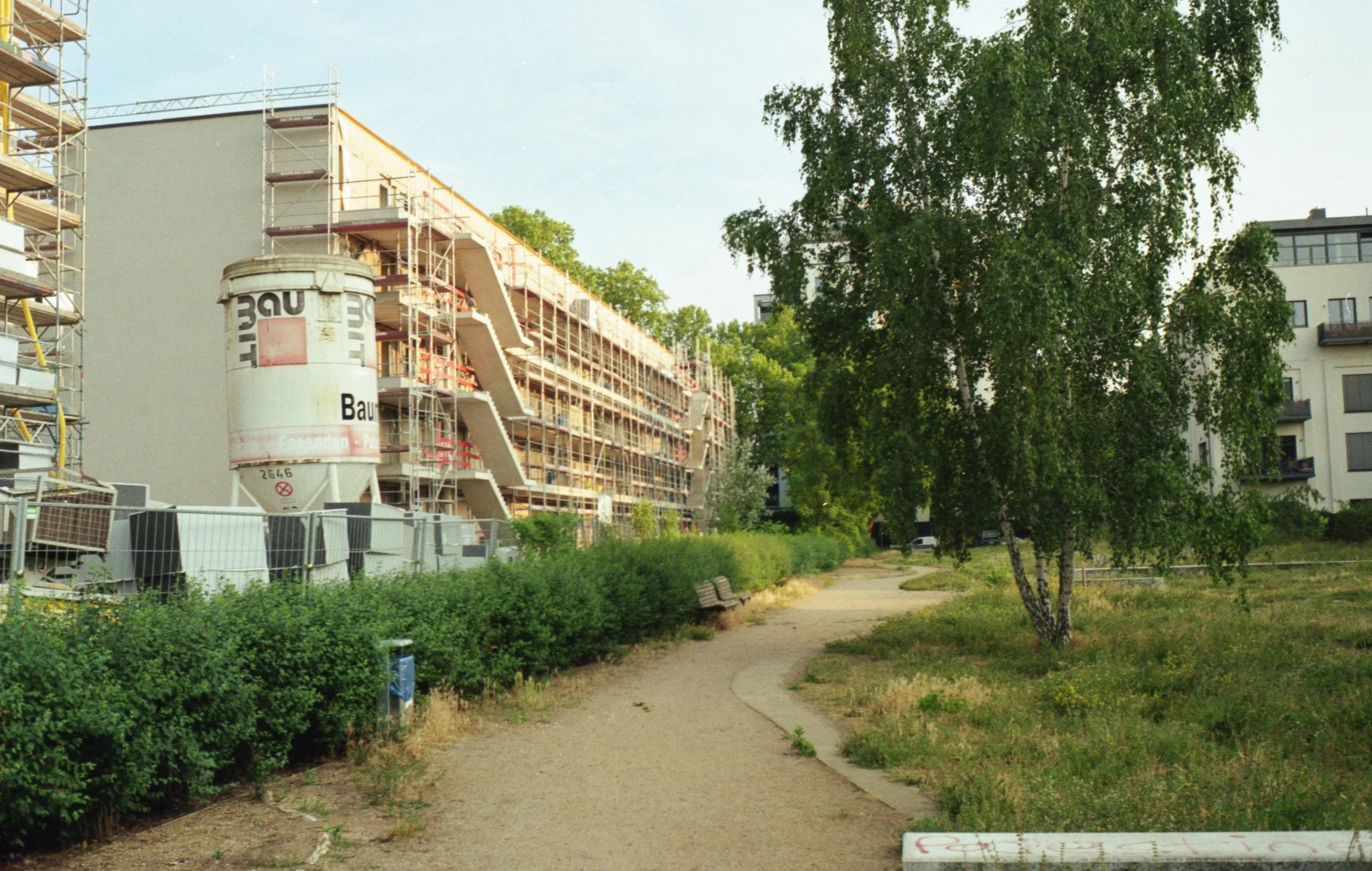 A daytime scene of a park with a winding dirt path, green bushes, and a large tree in the foreground. In the background is a modern, multi-story building and a construction site with scaffolding and a large white cement mixer.