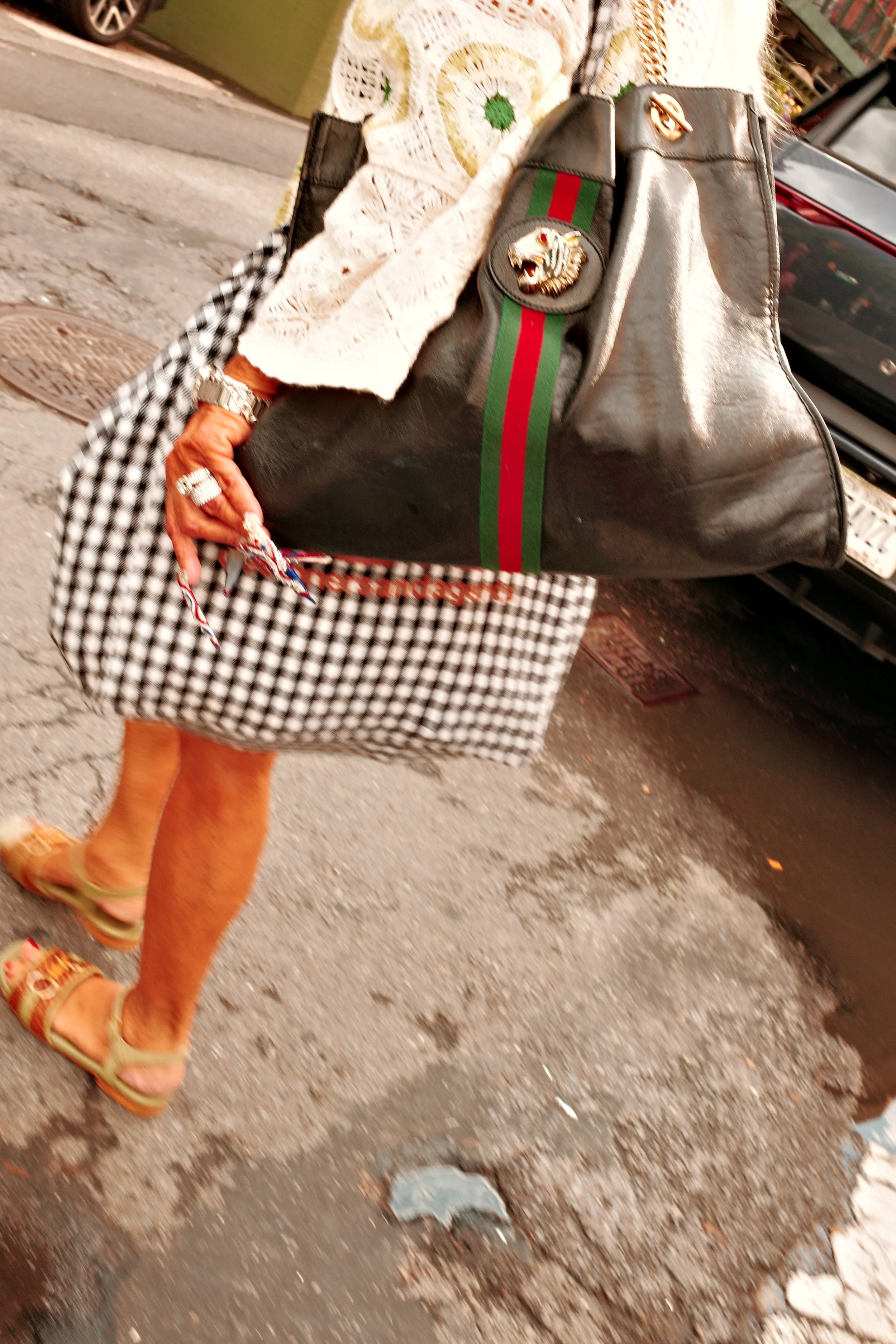Person walking on the sidewalk wearing sandals, plaid skirt, and jewelry, carrying a large black handbag with patches and a green-red stripe, with a black car and storefront in the background.