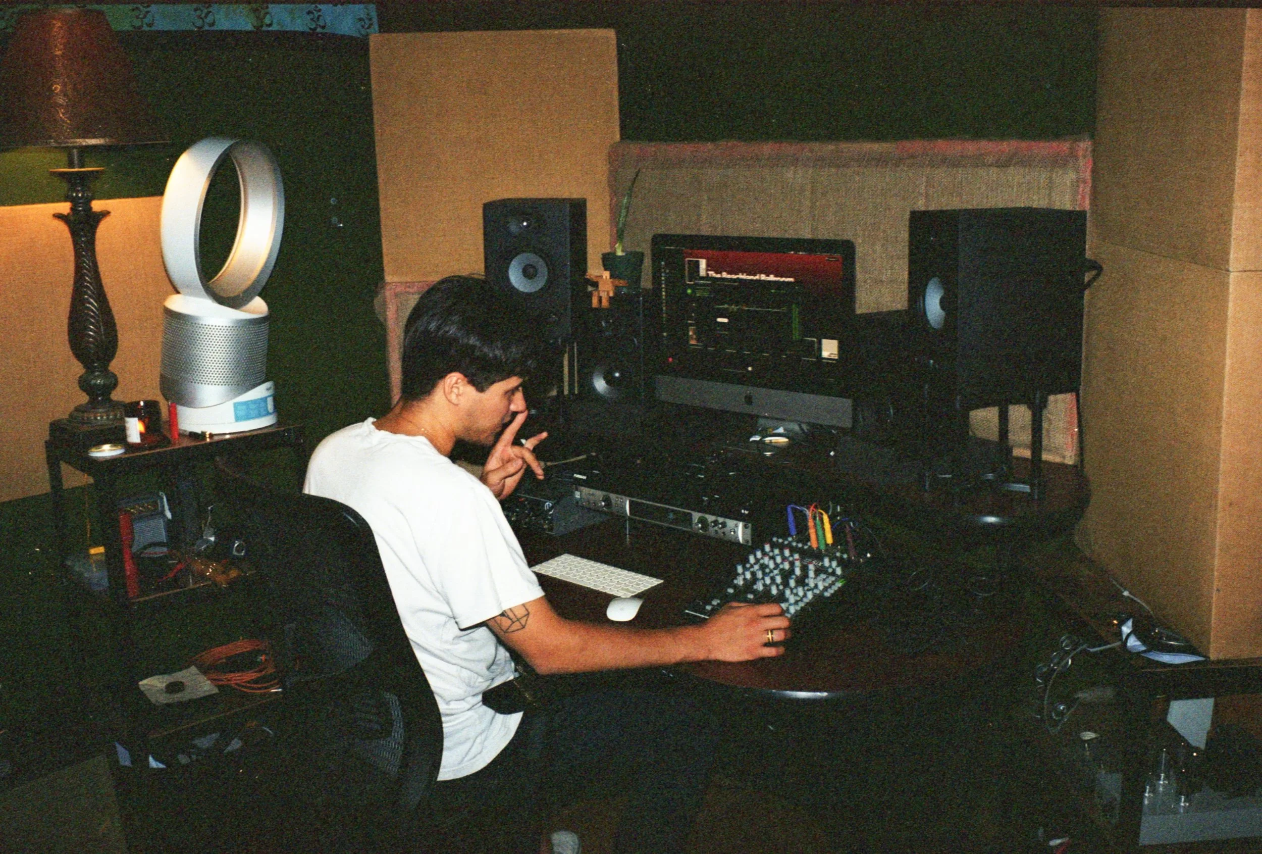 A person with dark hair and tattoos on their arm, wearing a white shirt, sits in front of a computer with audio equipment in a music production room. The room has a black desk, studio monitors, and soundproofing on the walls.