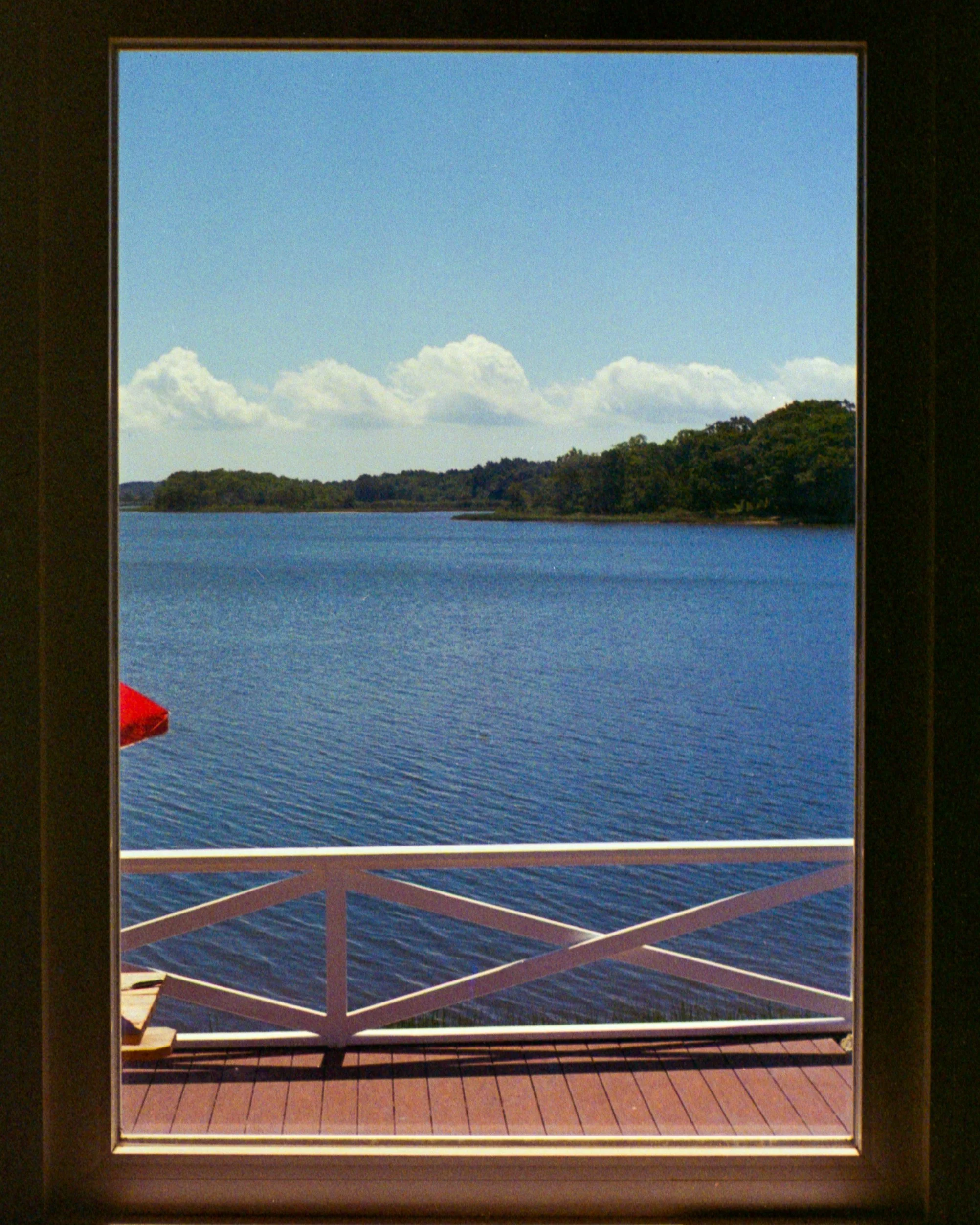 View of a lake or river with a shoreline covered with trees under a partly cloudy sky, seen through a window with a wooden railing and a wooden patio or balcony.