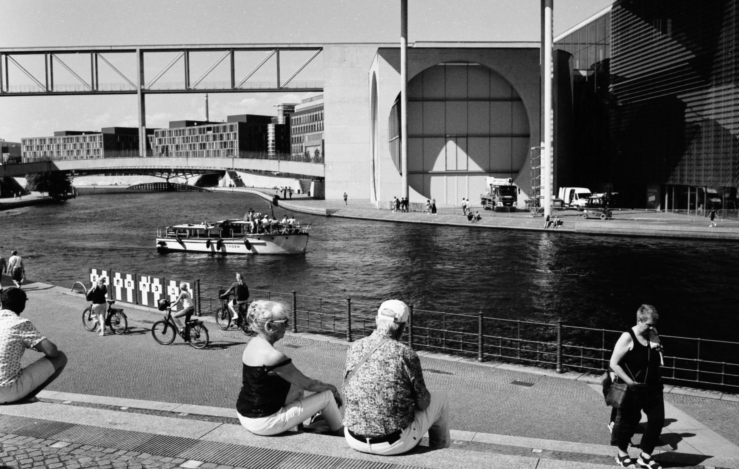 People sitting and biking along a riverside promenade with modern buildings and a boat in the water, with a bridge in the background.