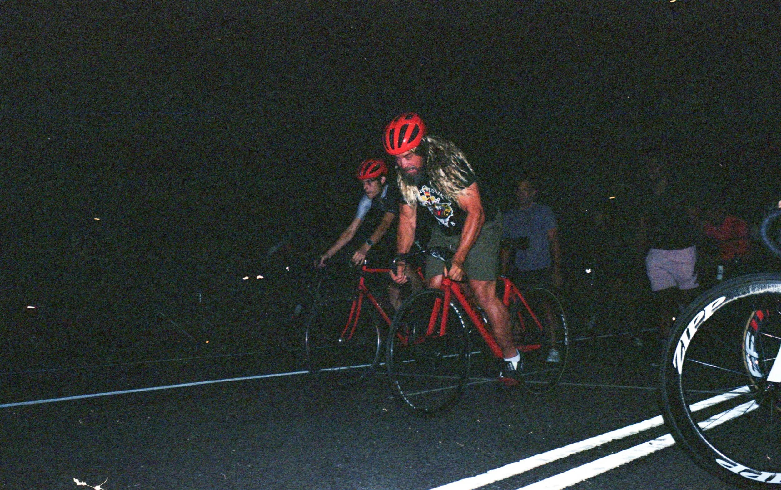 Nighttime image of a man with long hair and beard riding a red bike, wearing a black helmet and black cycling attire, with other cyclists and people in the background.