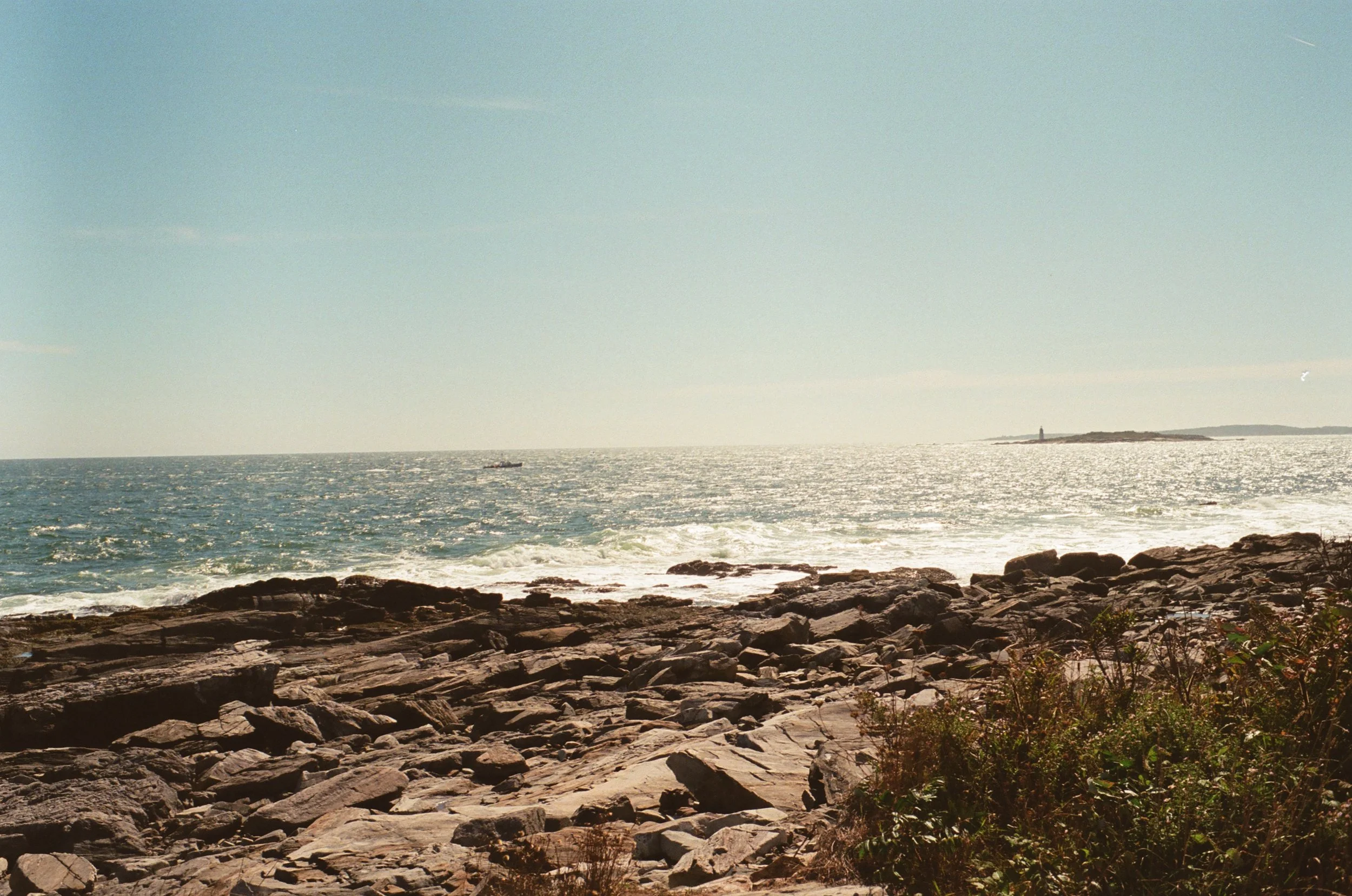 Rocky shoreline with the ocean in the background and a distant lighthouse on a small island.