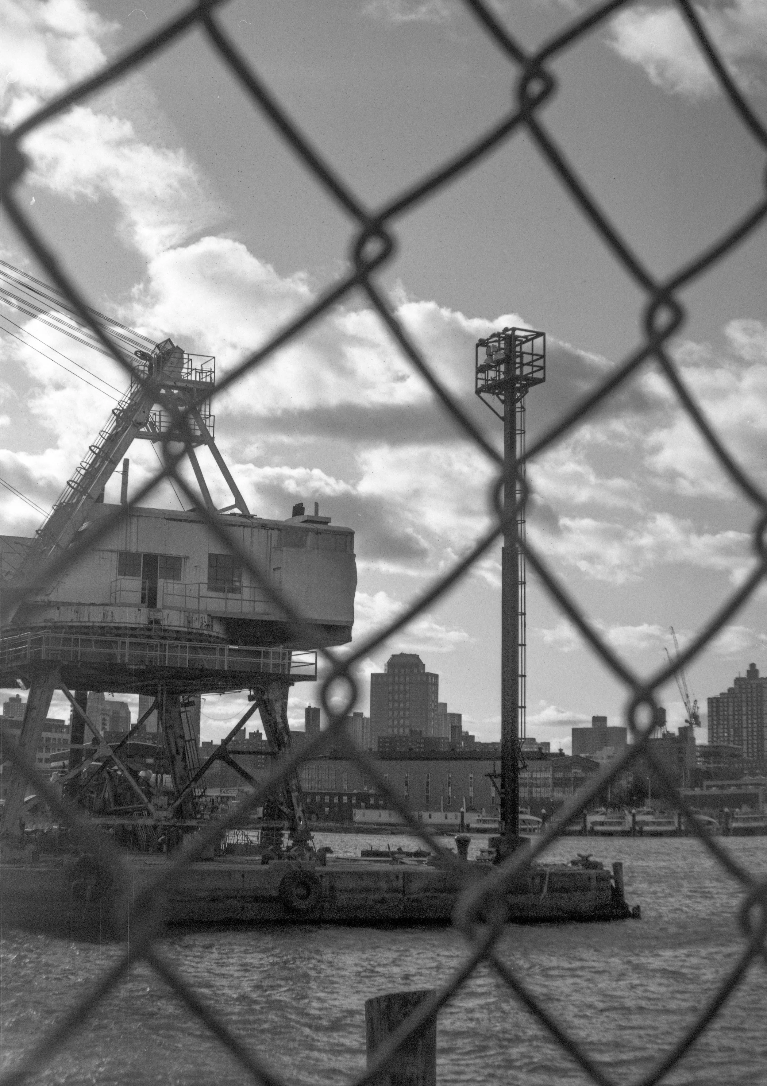Black and white photo showing a construction crane behind a chain-link fence, with city buildings in the background along a body of water.