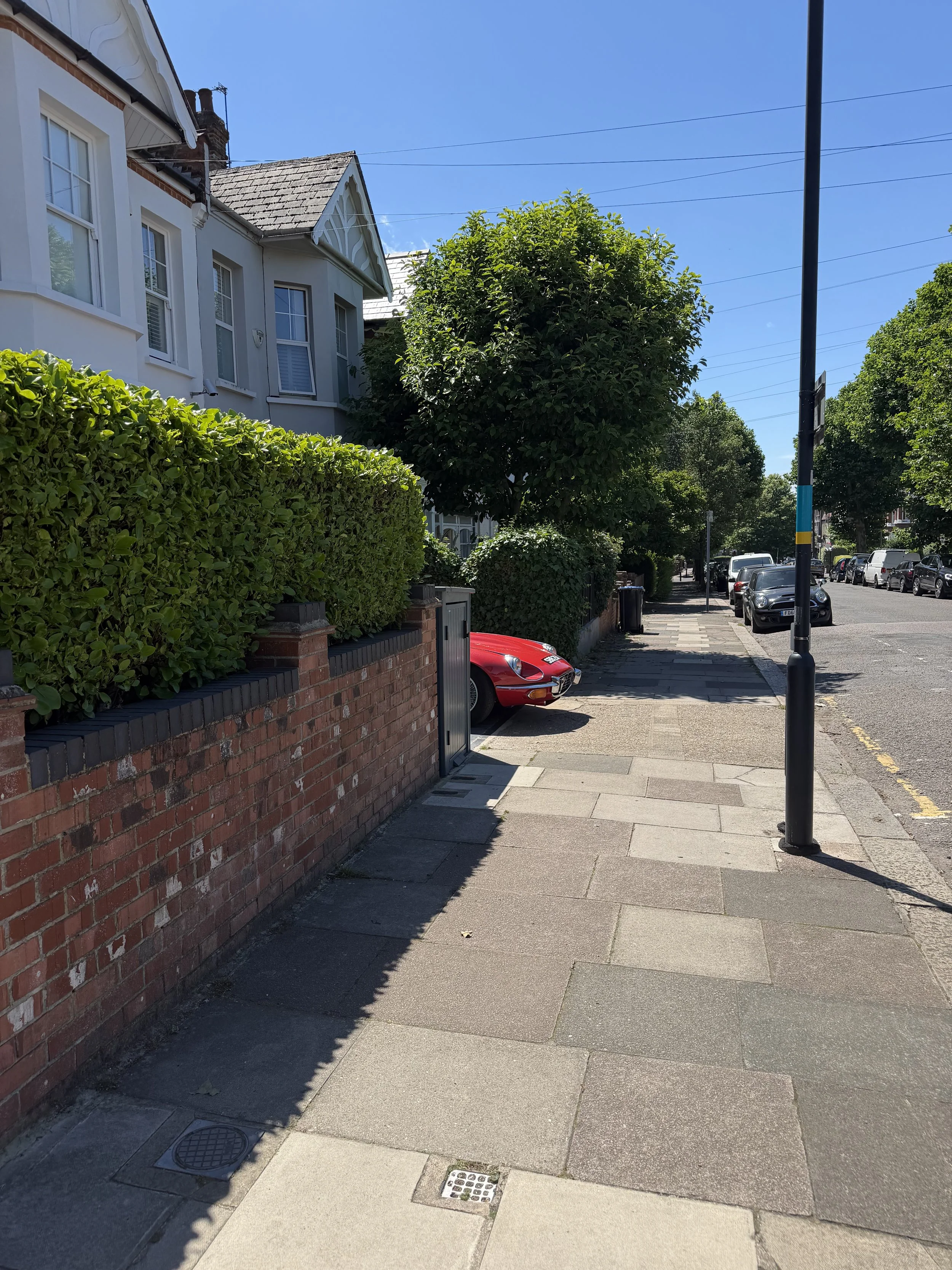Sidewalk with a brick wall, green hedge, trees, parked cars, a lamppost, and houses under a clear blue sky.