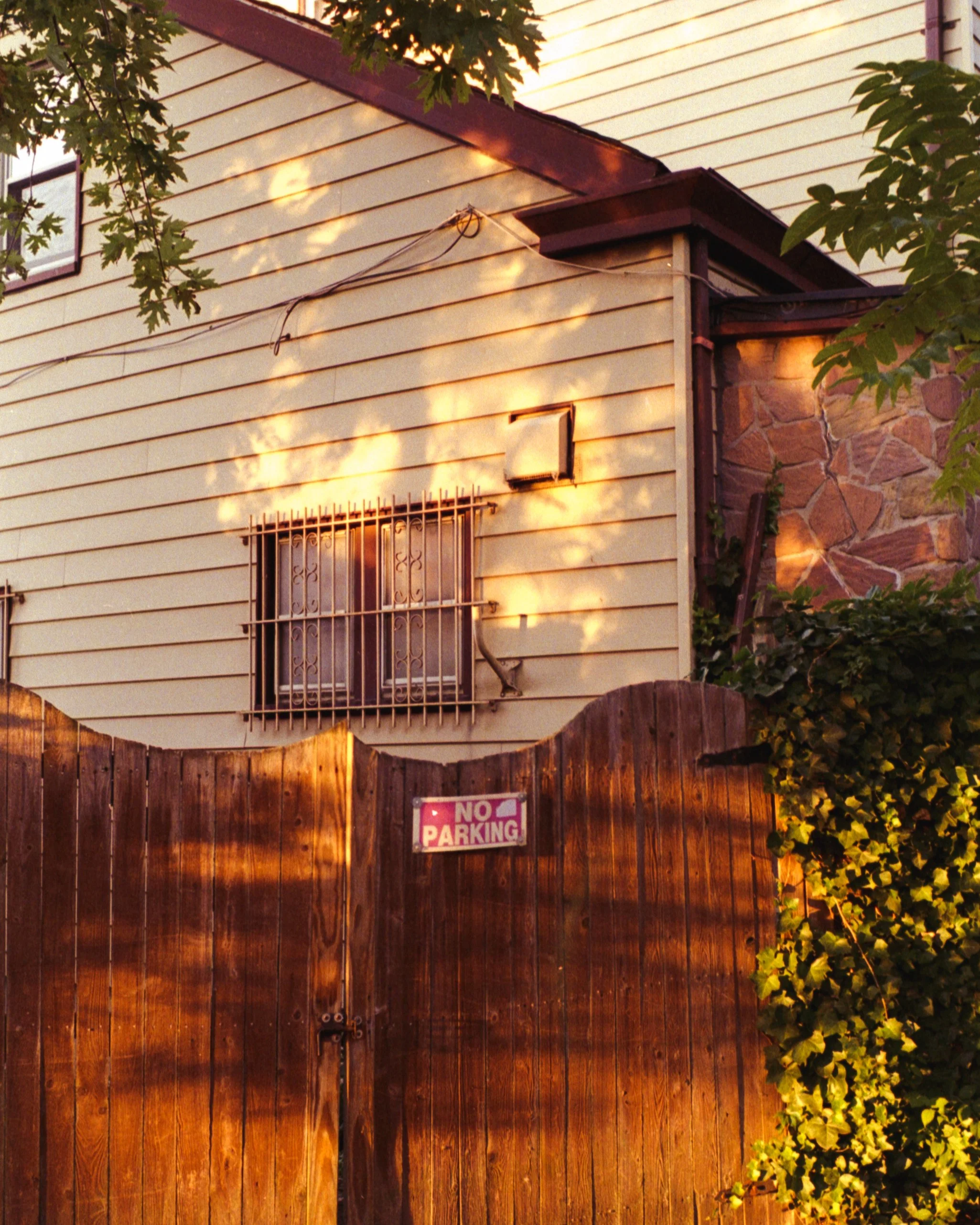 Wooden fence with a 'No Parking' sign, a house with yellow siding, barred window, and sandstone wall, leaves and trees in the foreground, and warm sunlight casting shadows.