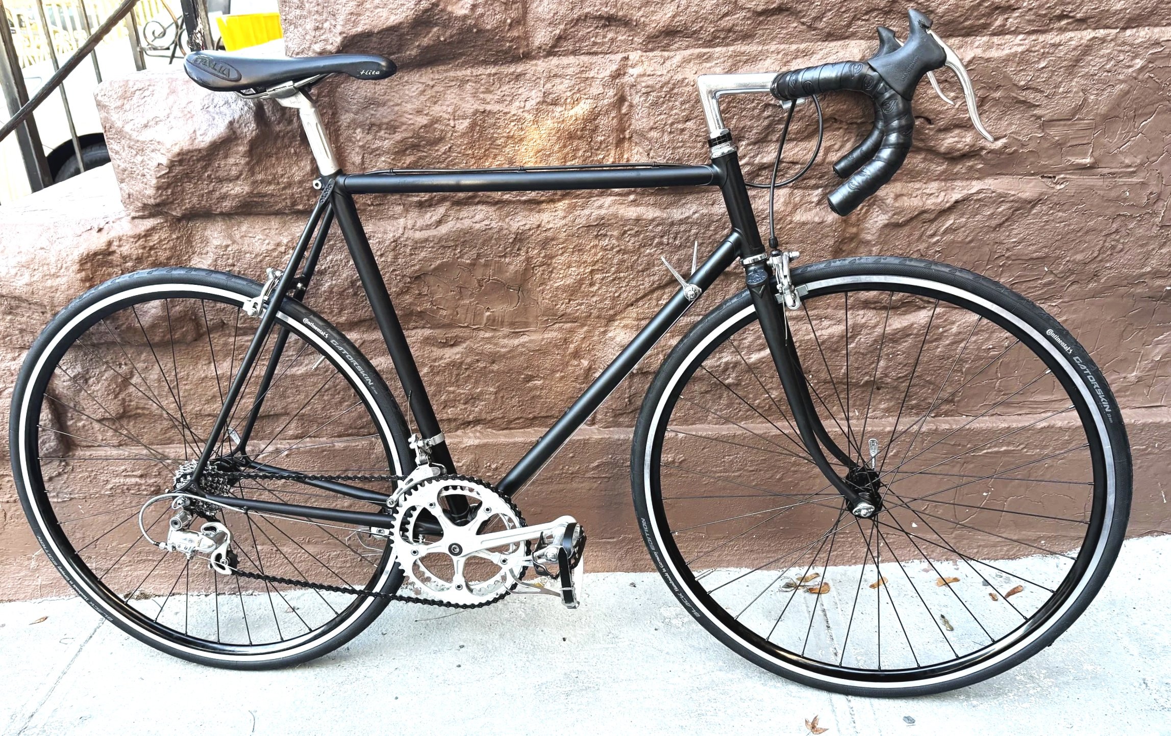 Black road bicycle leaning against a textured brown brick wall. Features include drop handlebars, a black saddle, thin tires, and silver gears and chain.