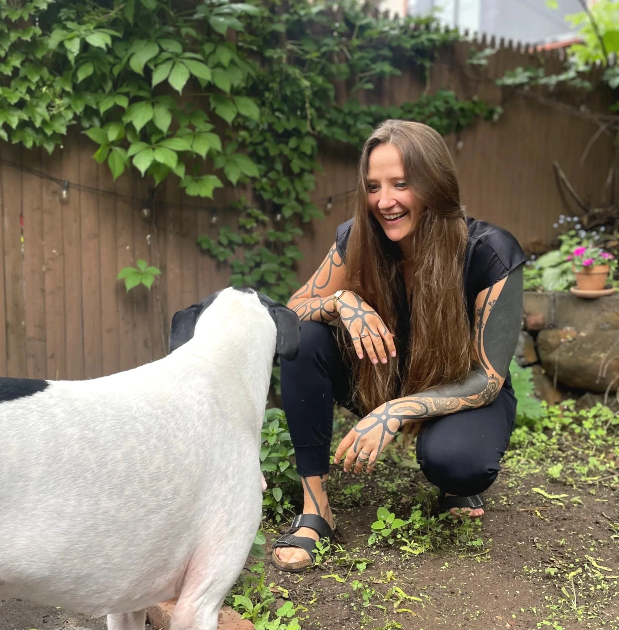 A woman with long brown hair, tattoos on her arms, black clothing, and sandals crouches in a garden, smiling at a white and black dog facing her.