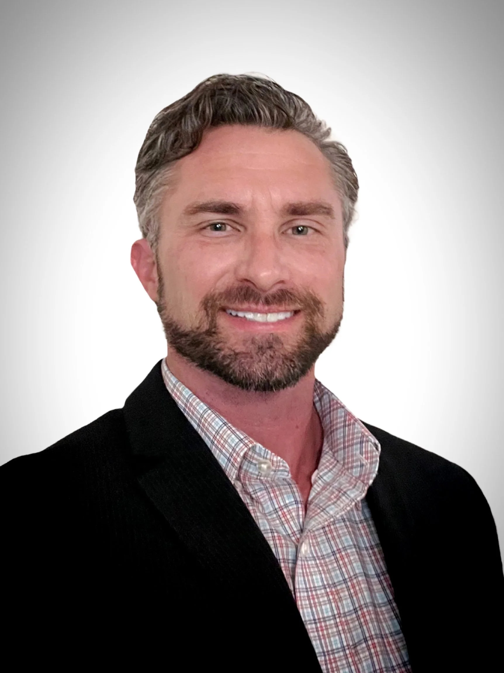 A headshot of a man with light skin, brown hair, and a beard, smiling, dressed in a checkered shirt and a dark blazer, against a plain background.