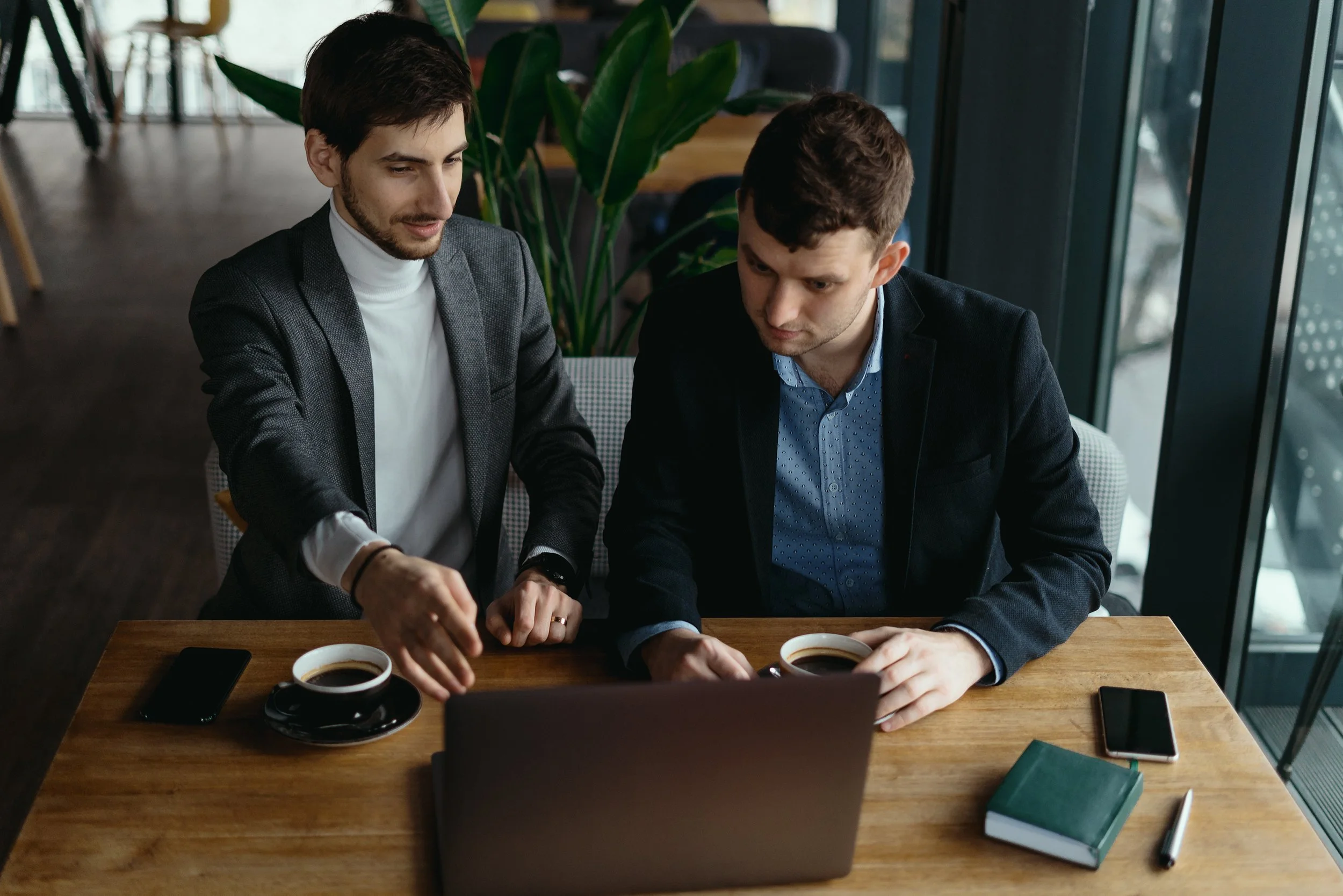 Two men in business attire working together at a wooden table with laptops, coffee cups, a notebook, and pens in a modern office.