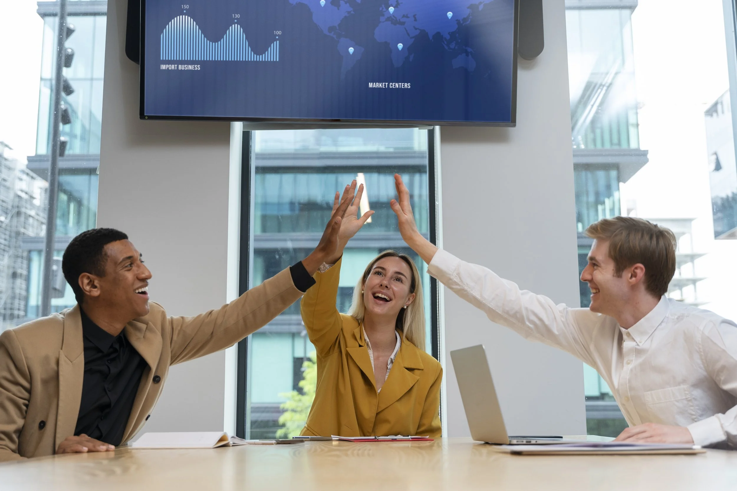 three colleagues giving each other a high five in a modern office with a city view, a monitor displaying a world map and charts in the background