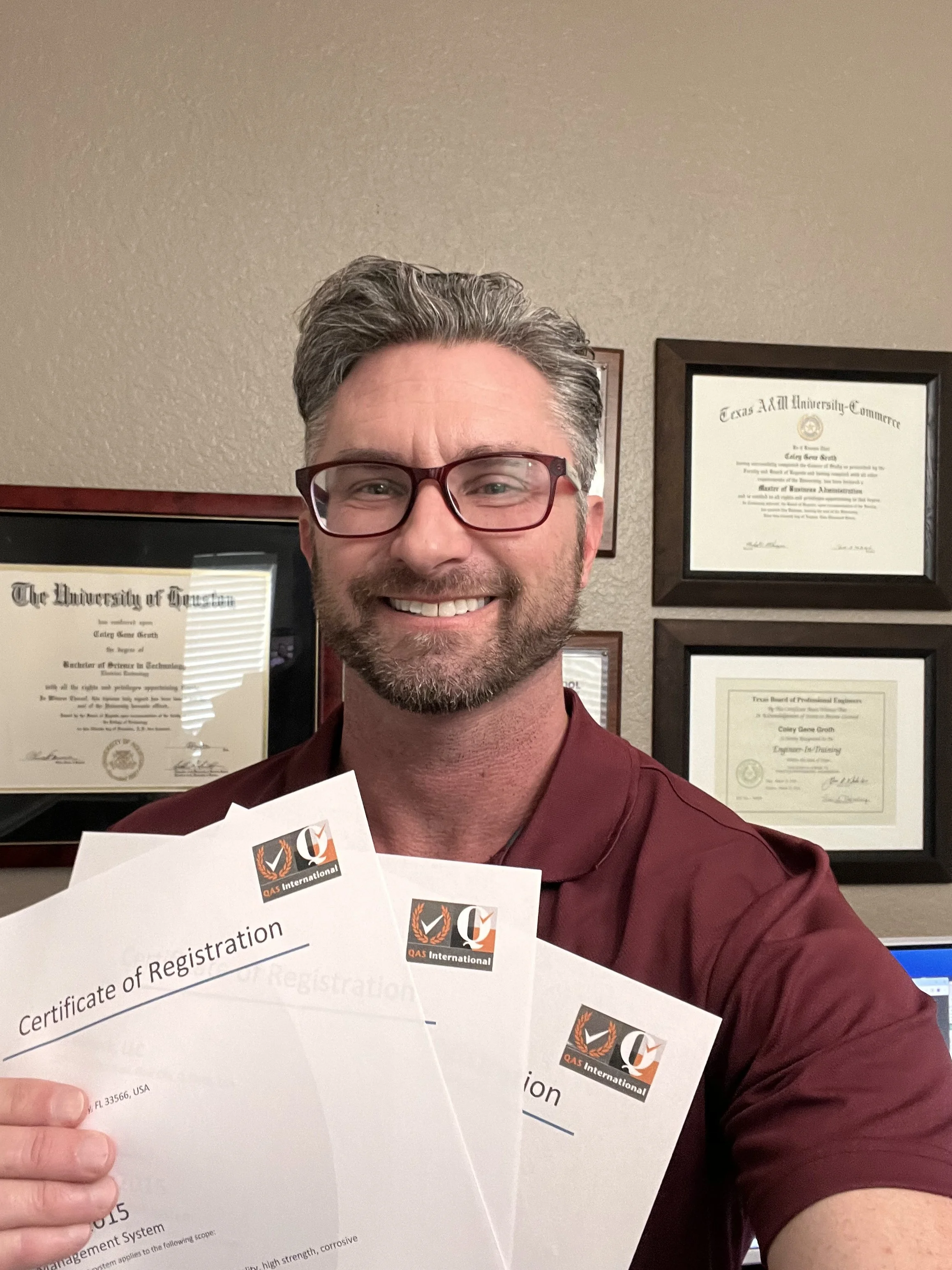A man with glasses and a beard smiling, holding three certificates from QAS International in front of a wall with framed diplomas and certifications.