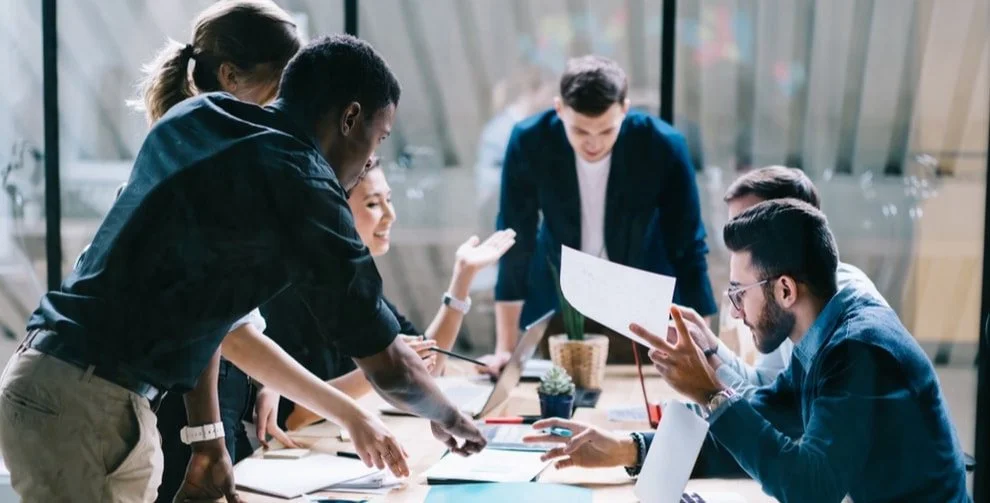A diverse group of colleagues in a modern office engaging in a lively discussion around a table filled with laptops and documents.