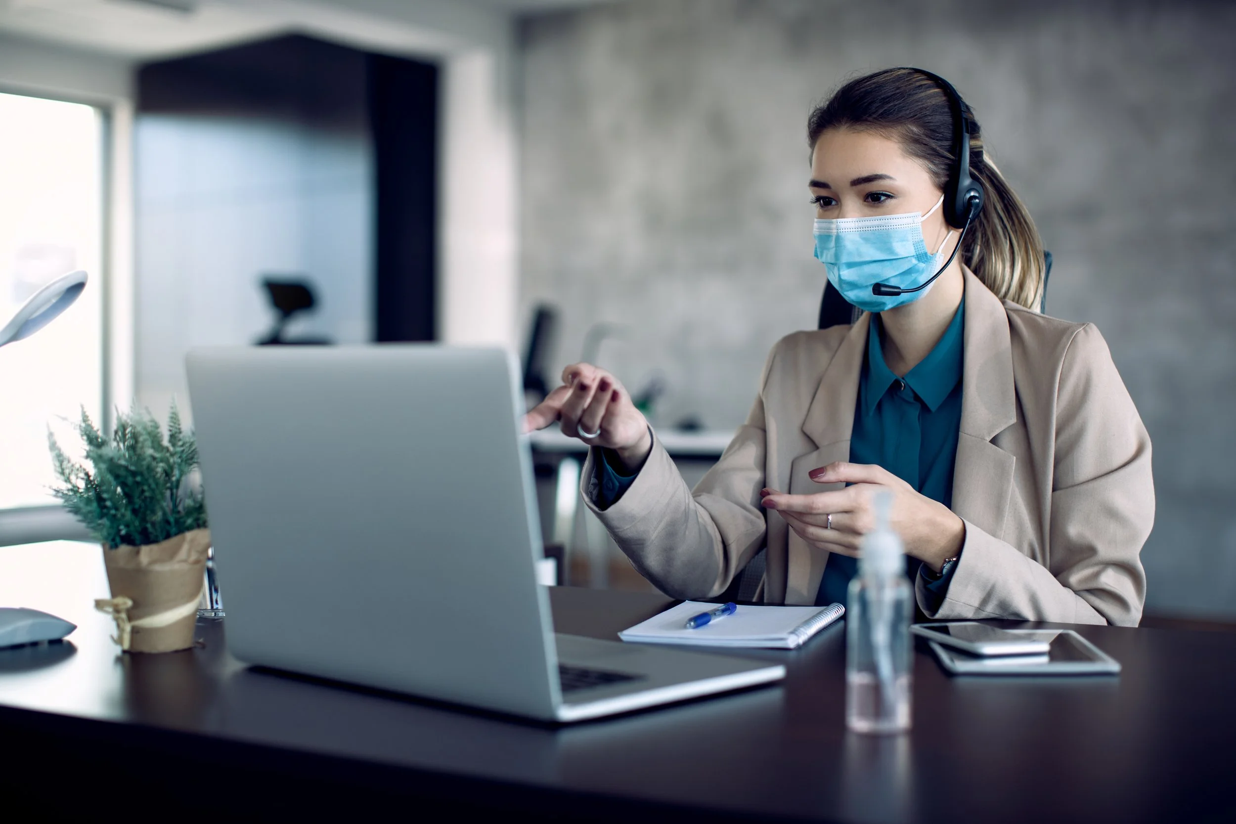 A woman wearing a face mask and headset working at her desk with a laptop, notebook, tablet, and a hand sanitizer bottle in a modern office.